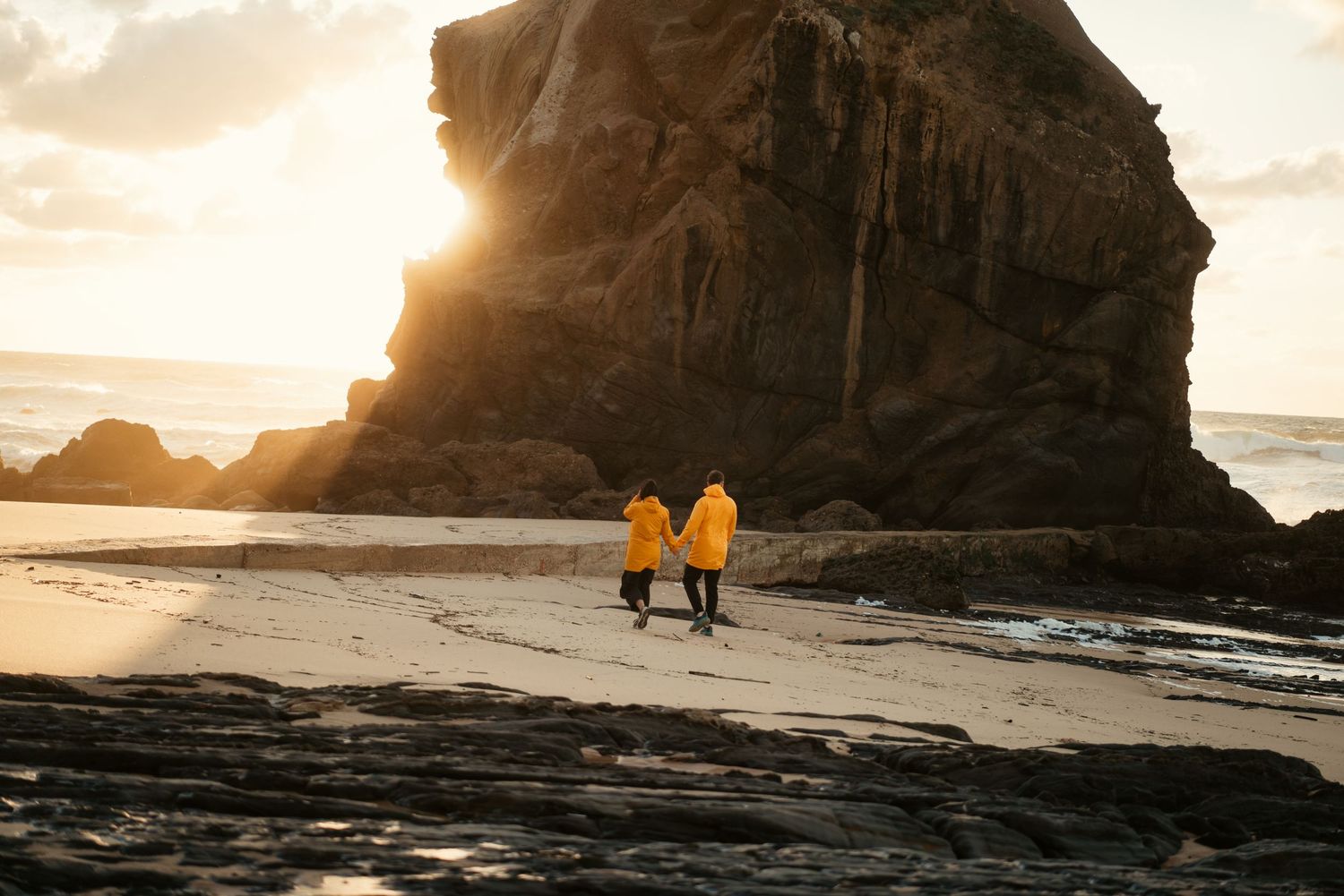 A couple in matching yellow raincoats walking on Santa Cruz beach at golden hour, couples photography Portugal