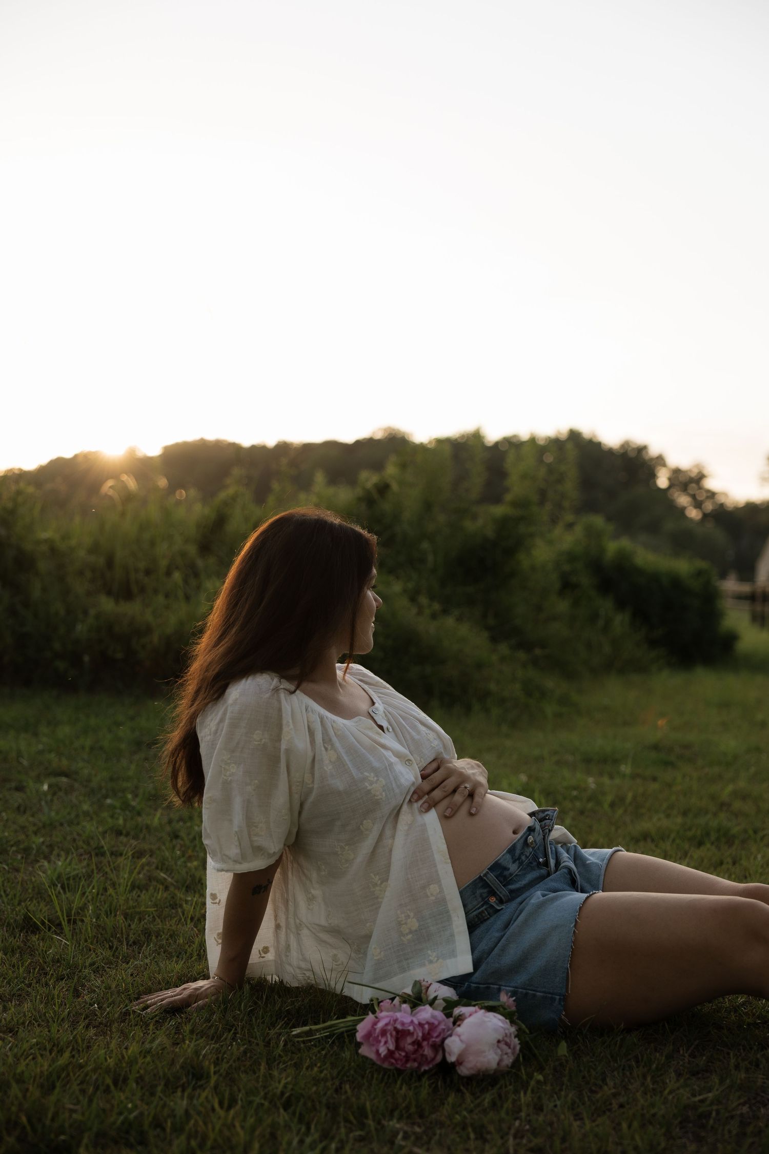 Pregnant woman in white top and denim shorts sitting on grass at sunset, holding belly with pink peonies nearby.