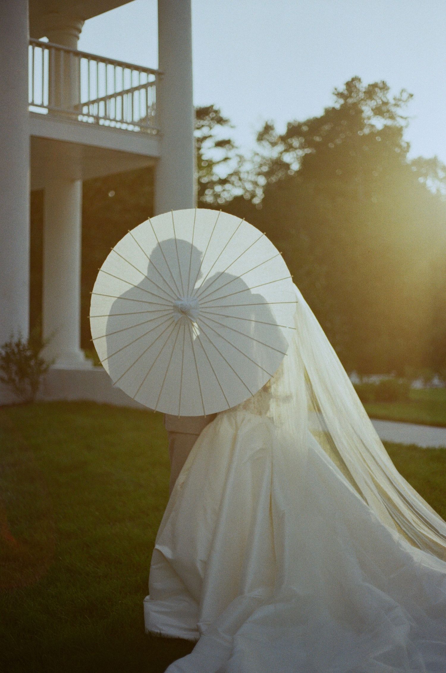 Silhouette of a couple kissing behind a white paper parasol at golden hour, the bride's gown and veil flowing near a columned porch.