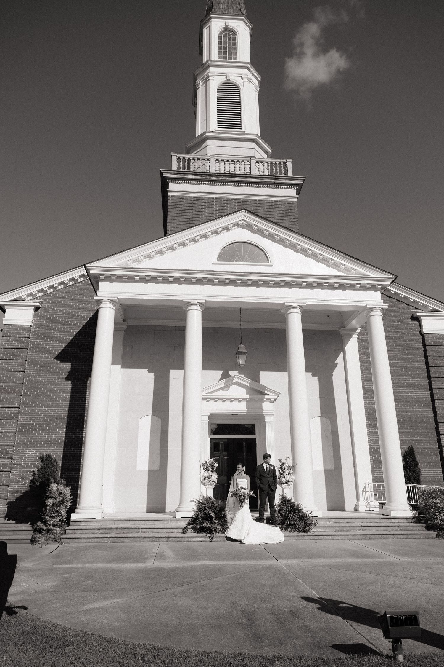Black and white photo of a bride and groom standing before a classic white-columned church with a tall steeple.