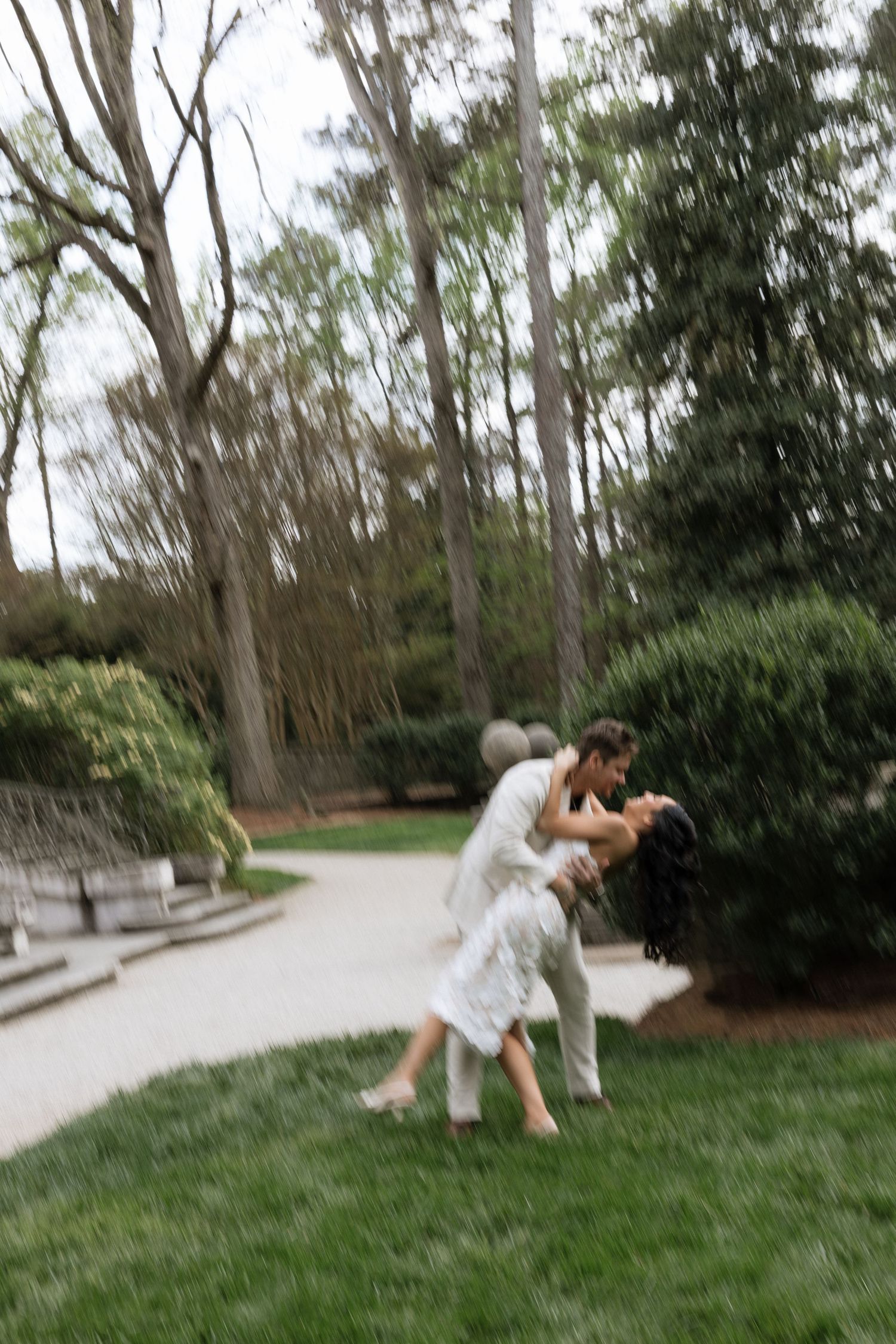 Couple sharing a romantic dip kiss in a lush green park garden surrounded by tall trees on a rainy day.