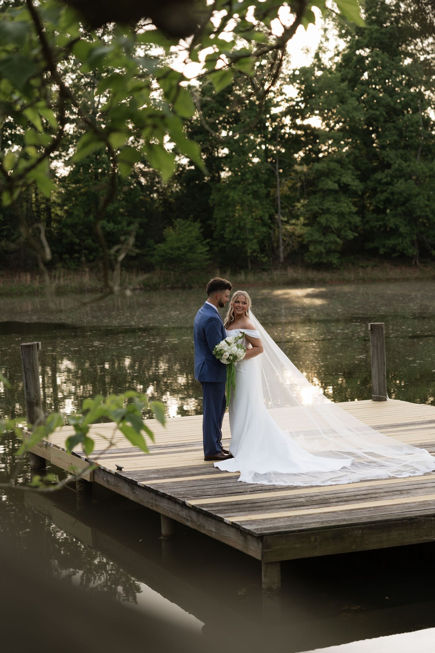 Bride and groom standing on a wooden dock by a serene lake surrounded by lush green trees during golden hour.