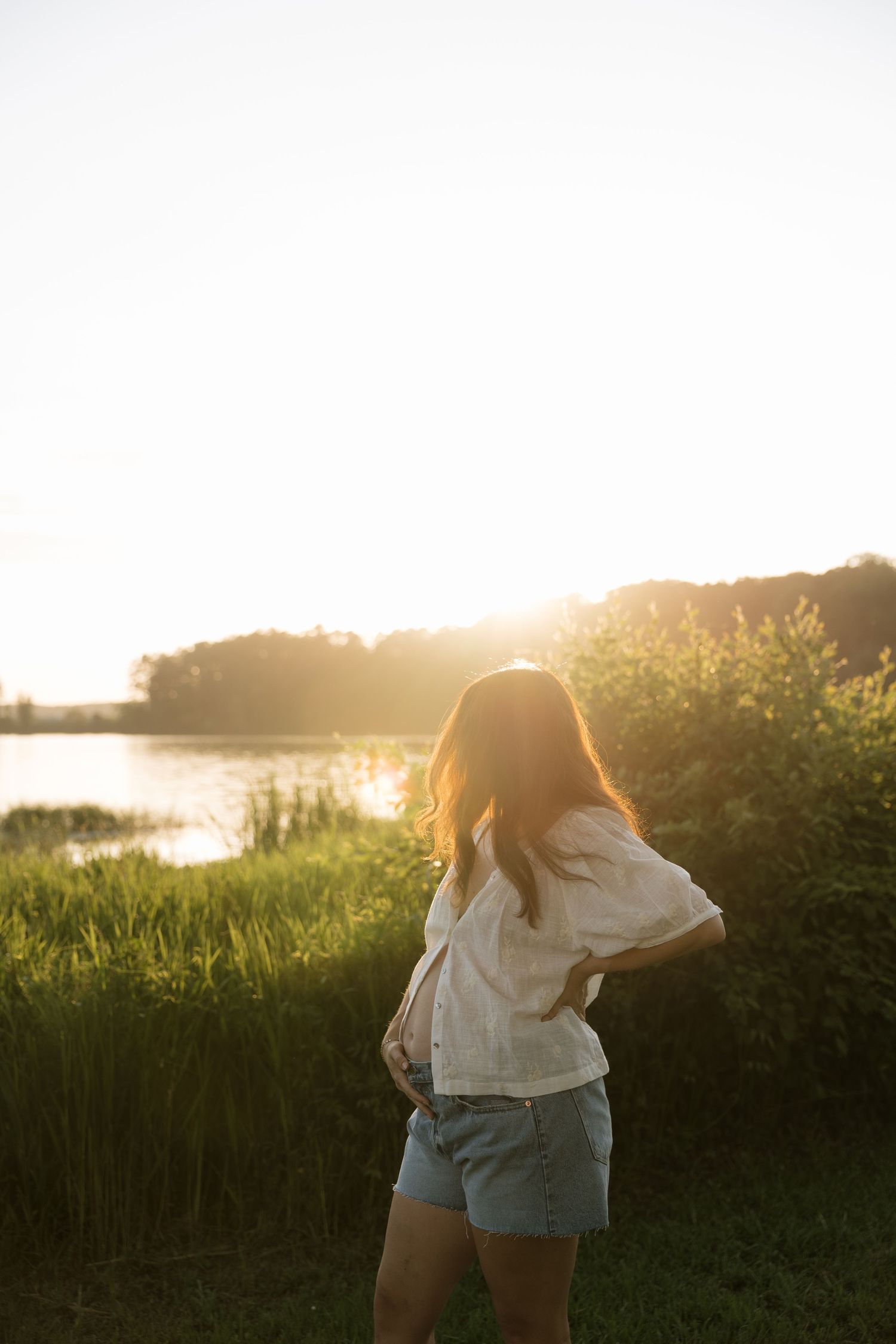 Woman in white blouse and denim shorts standing by a lake at golden sunset with lush green grass and trees.