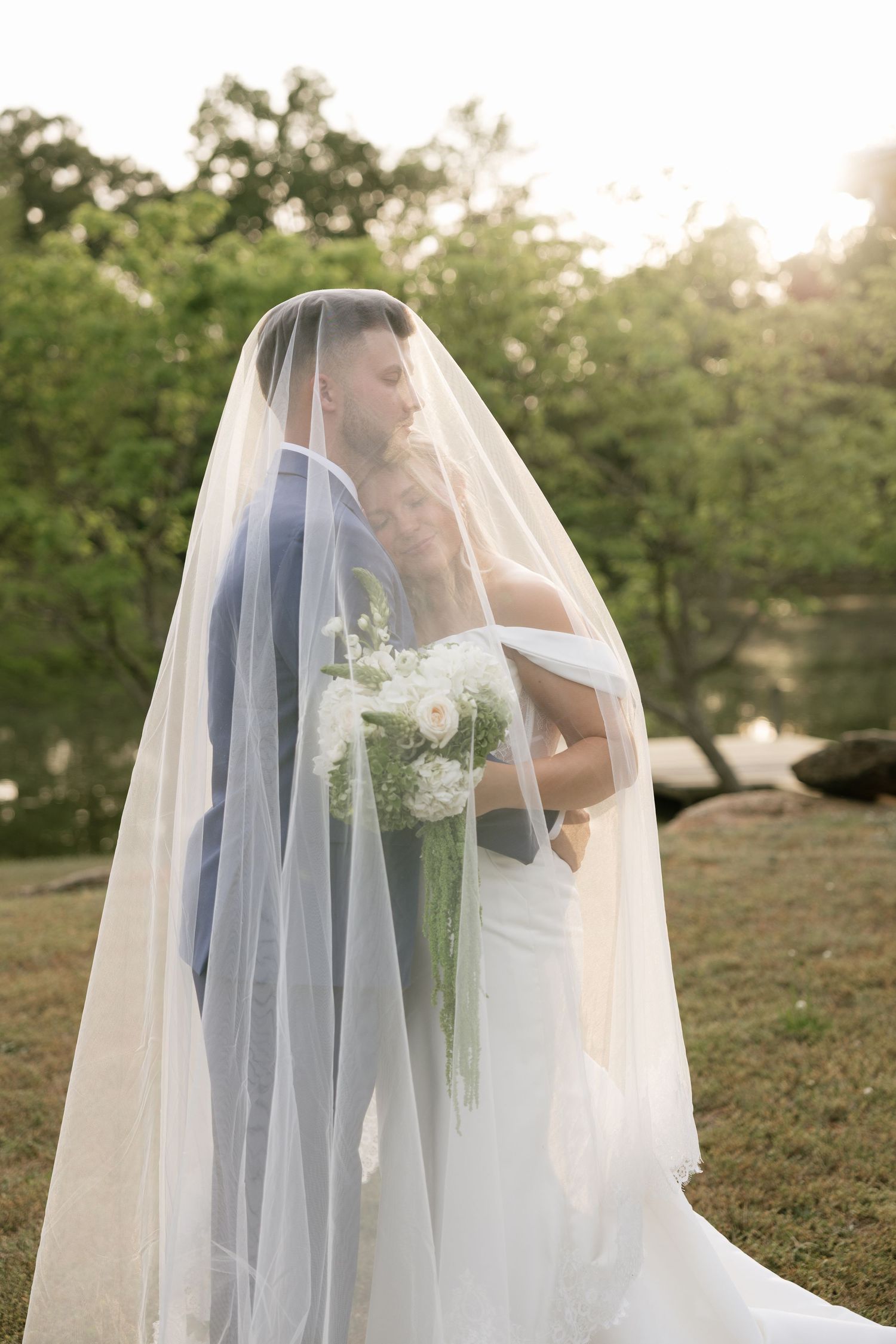 Bride and groom sharing a romantic kiss under a flowing white veil outdoors, surrounded by lush greenery.
