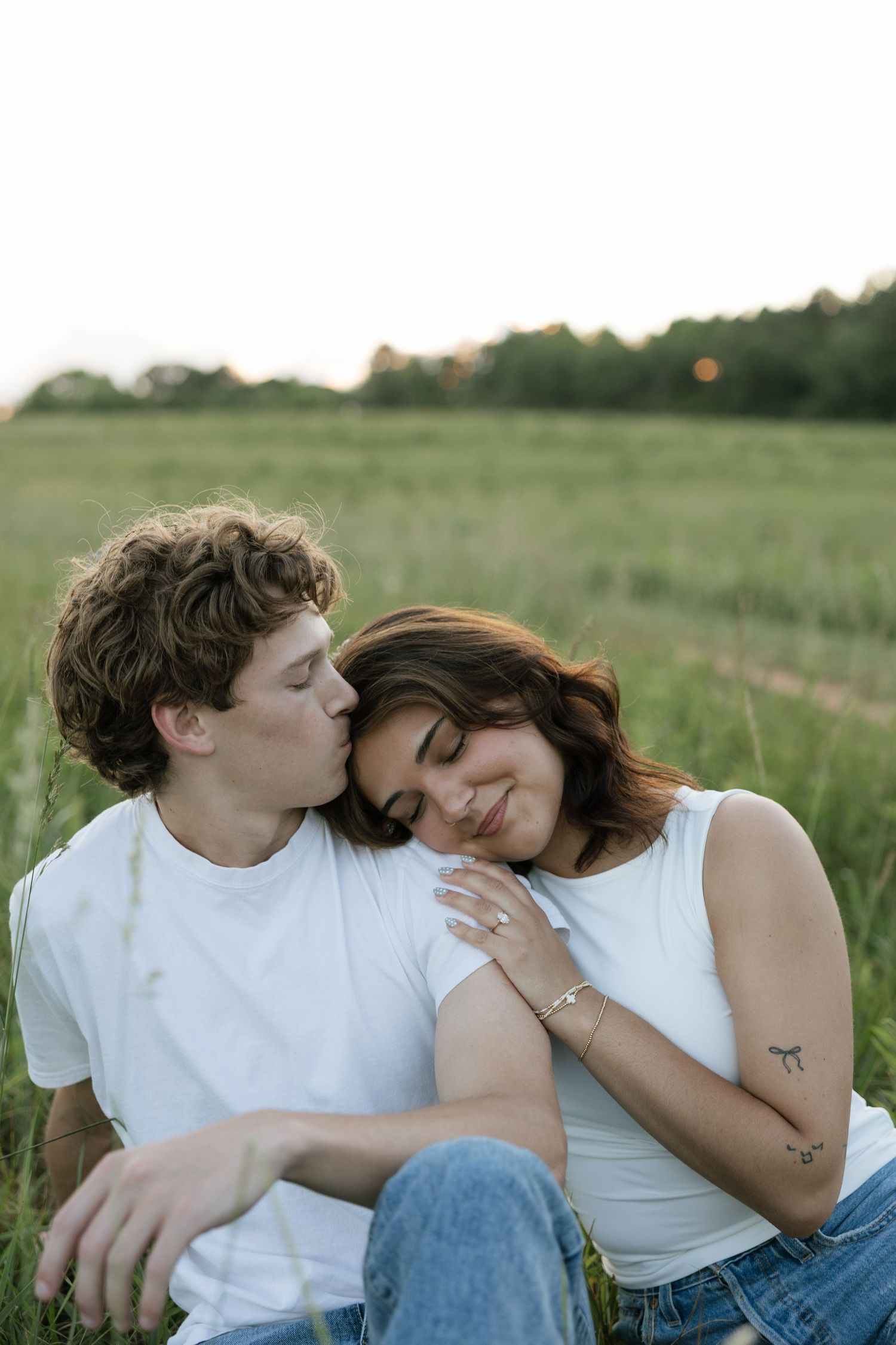 A young couple sits in a green field, sharing a tender moment as he kisses her cheek at sunset.