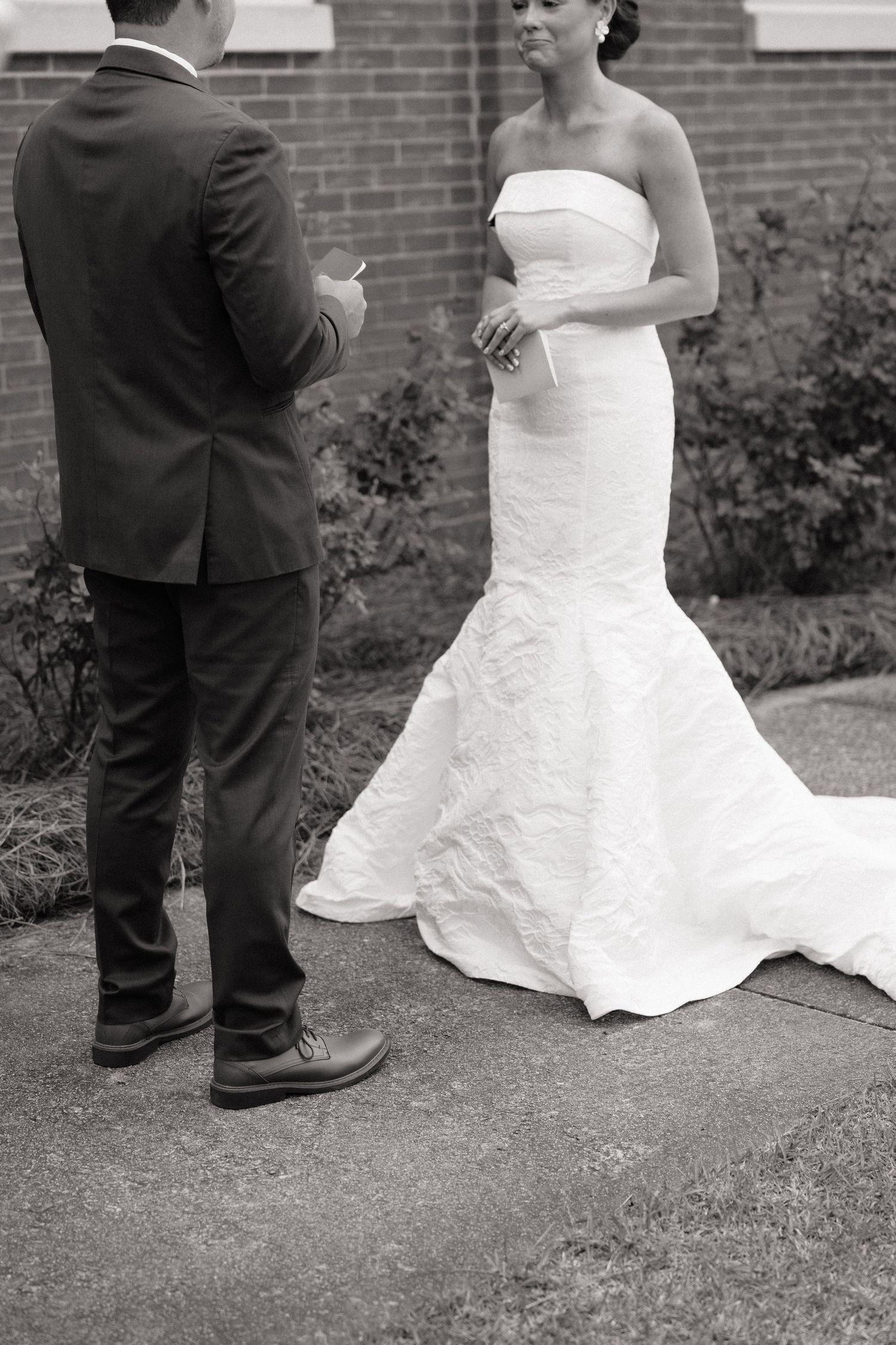 Black and white photo of a bride in a strapless mermaid gown and groom exchanging vows outdoors.