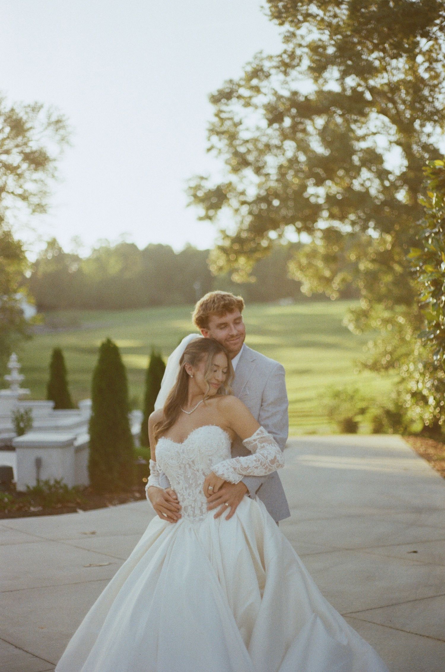 Groom embracing bride from behind in a satin gown with lace sleeves on a sunlit garden path overlooking a green lawn.