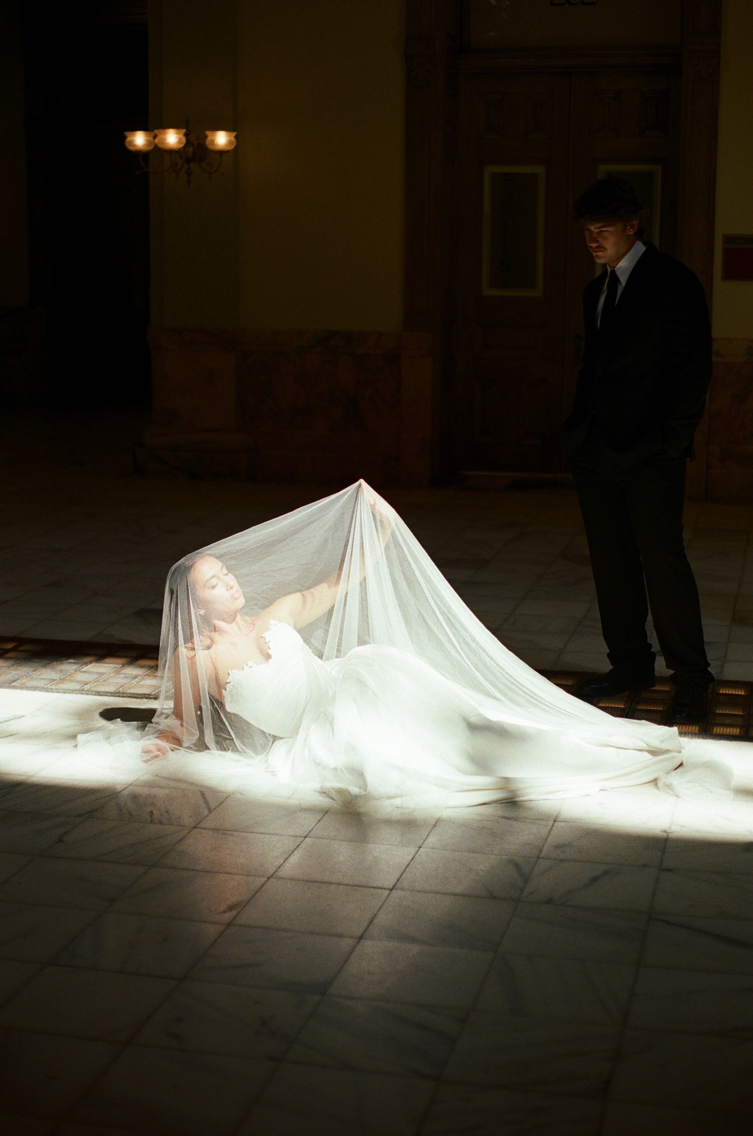 A bride in a white gown reclines on a tiled floor beneath a sheer veil in a beam of light as a man in a dark suit watches.