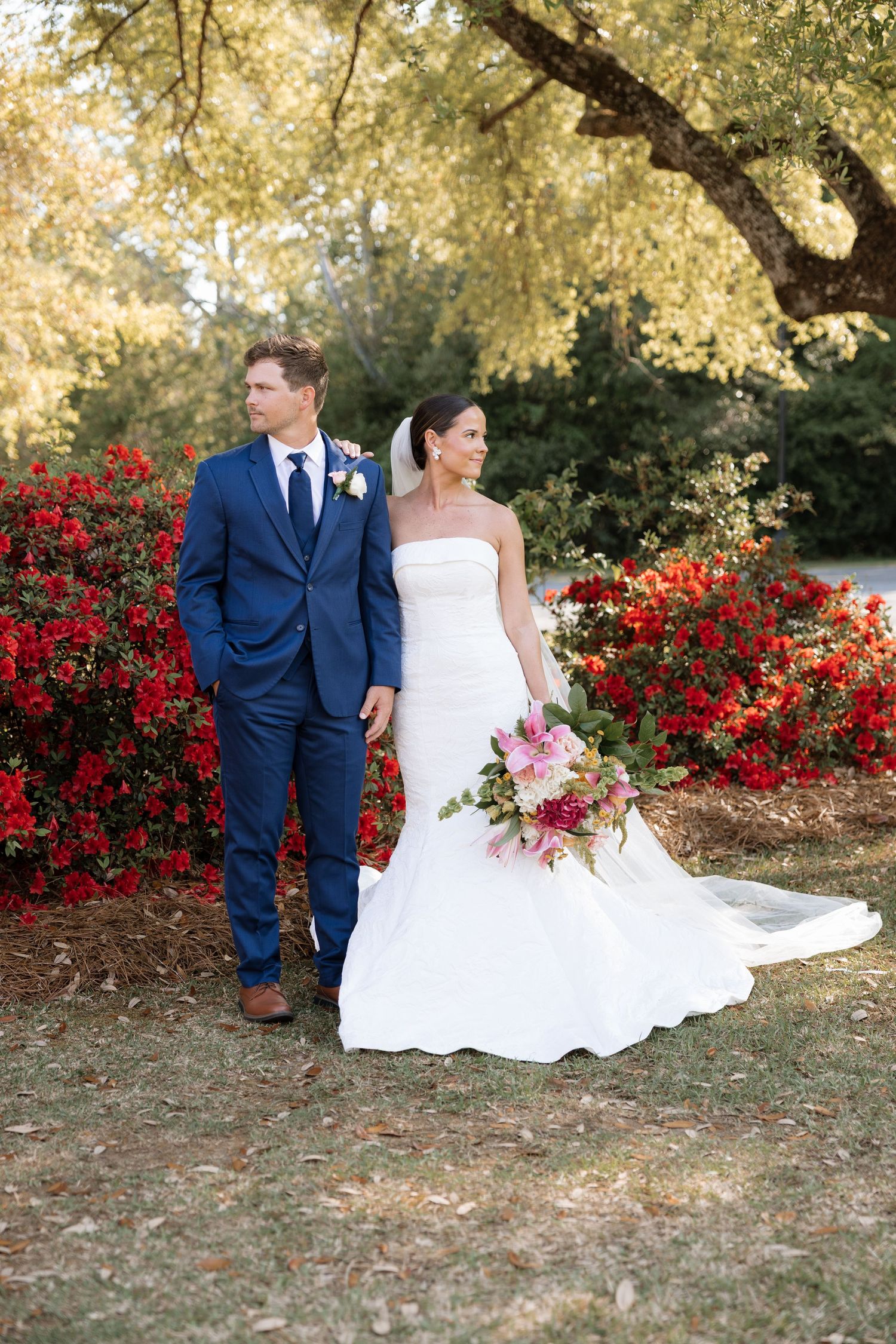 Bride and groom pose outdoors near vibrant red flowers, she holds a pink bouquet in a white gown, he wears a navy suit.