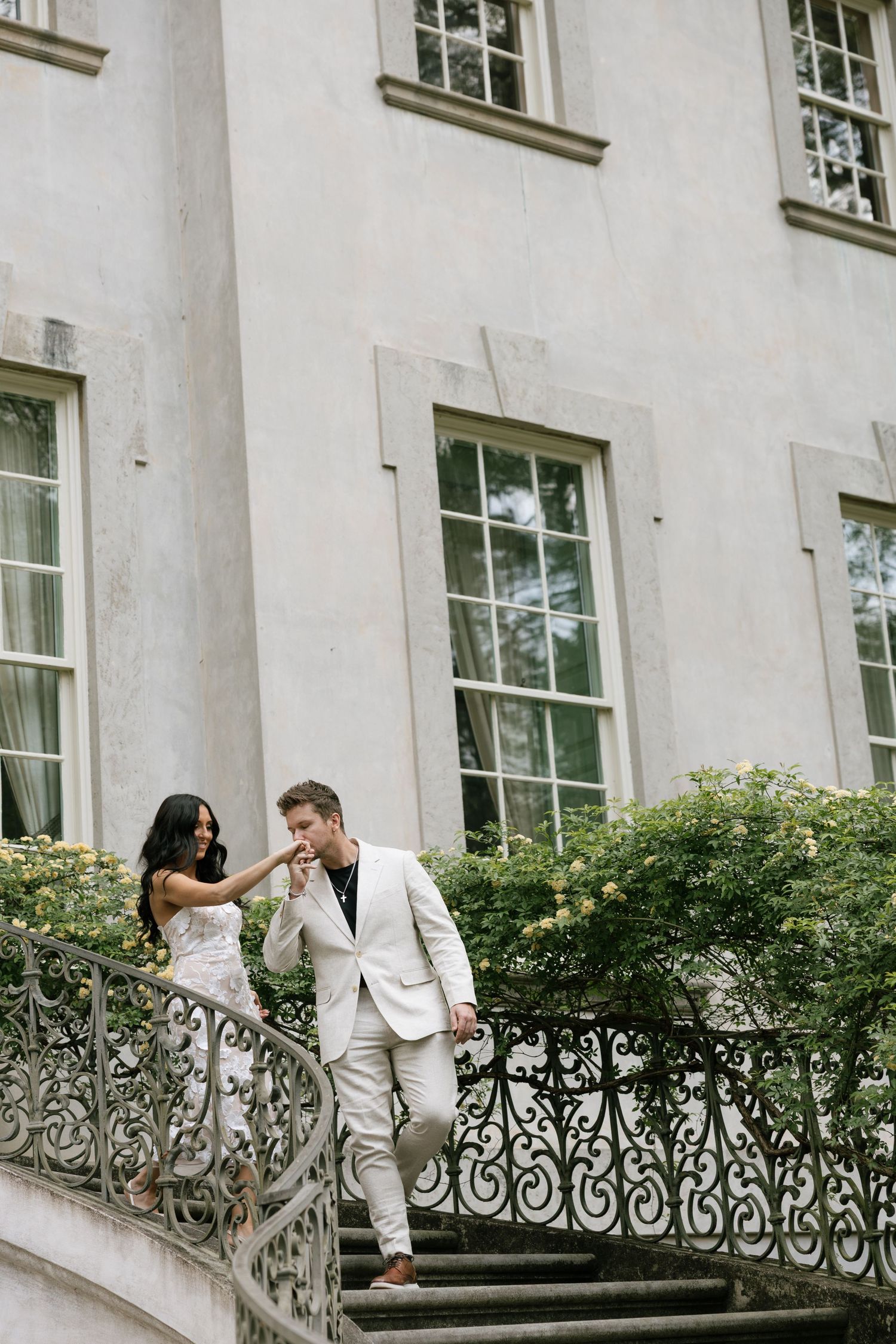 Elegant couple in white attire descending ornate iron staircase outside a grand estate building.