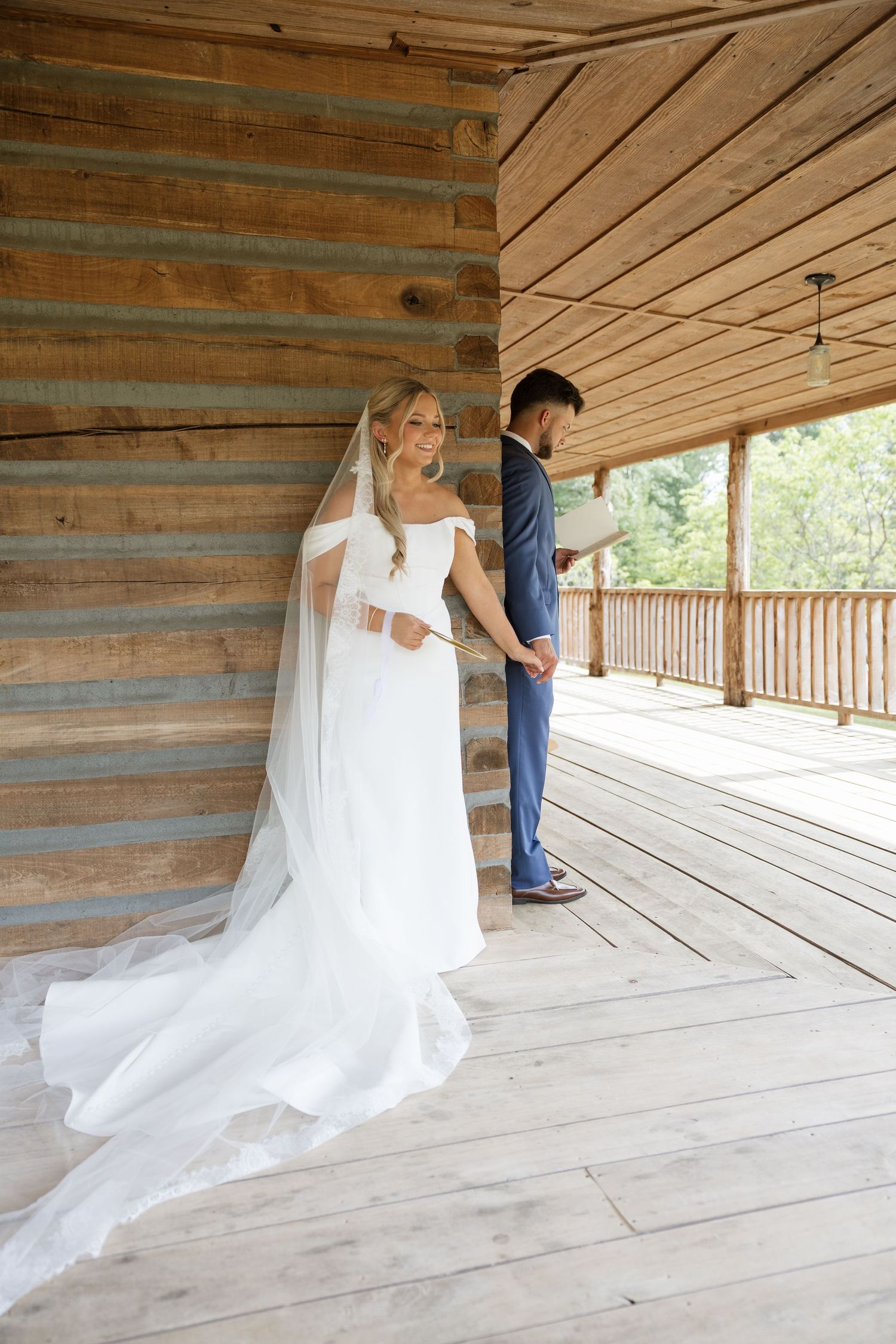 Bride in white gown and veil holds groom's hand on rustic wooden cabin porch during outdoor wedding ceremony.