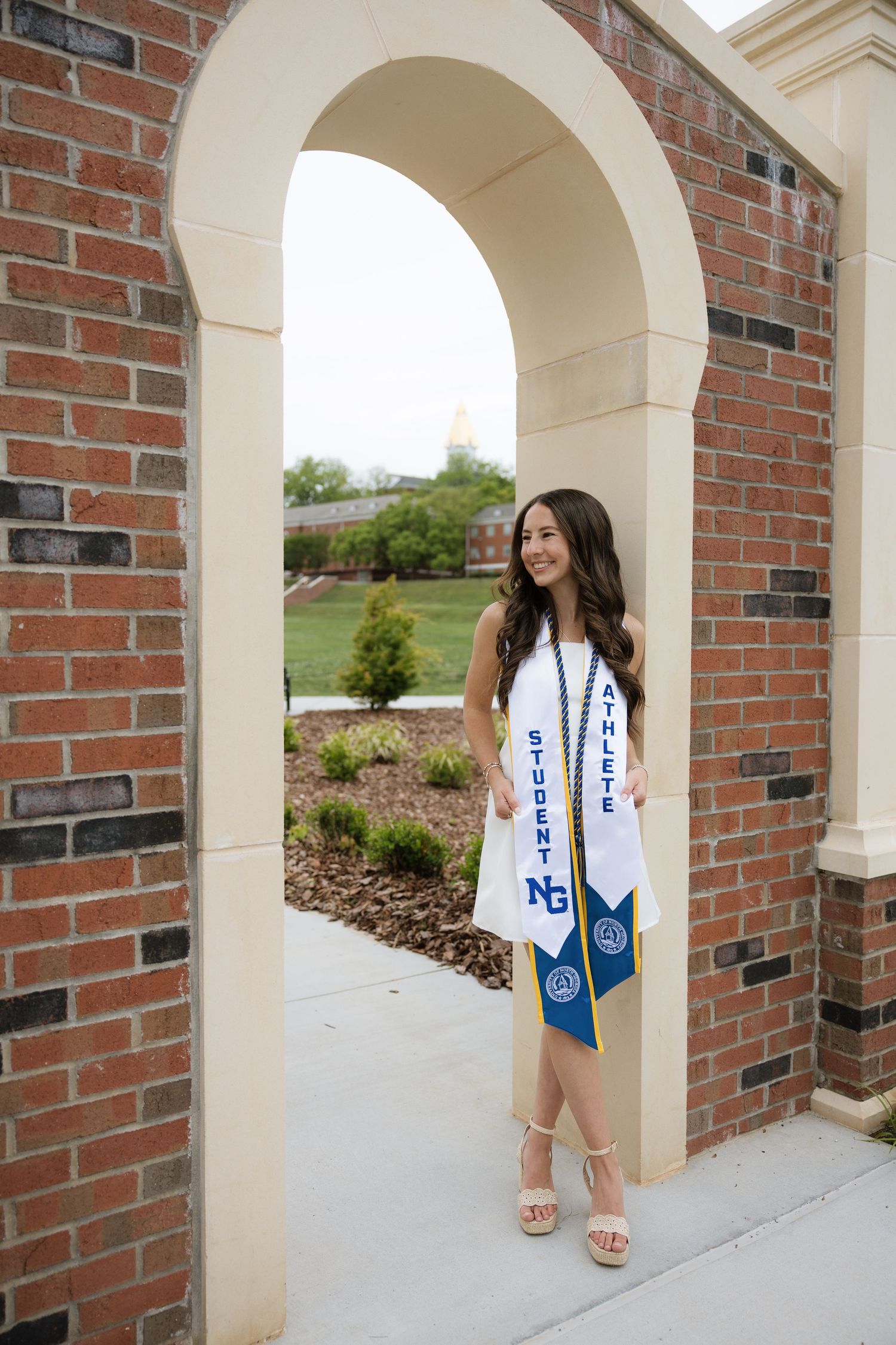 Female student-athlete poses in brick archway wearing graduation stole with NG logo outdoors.
