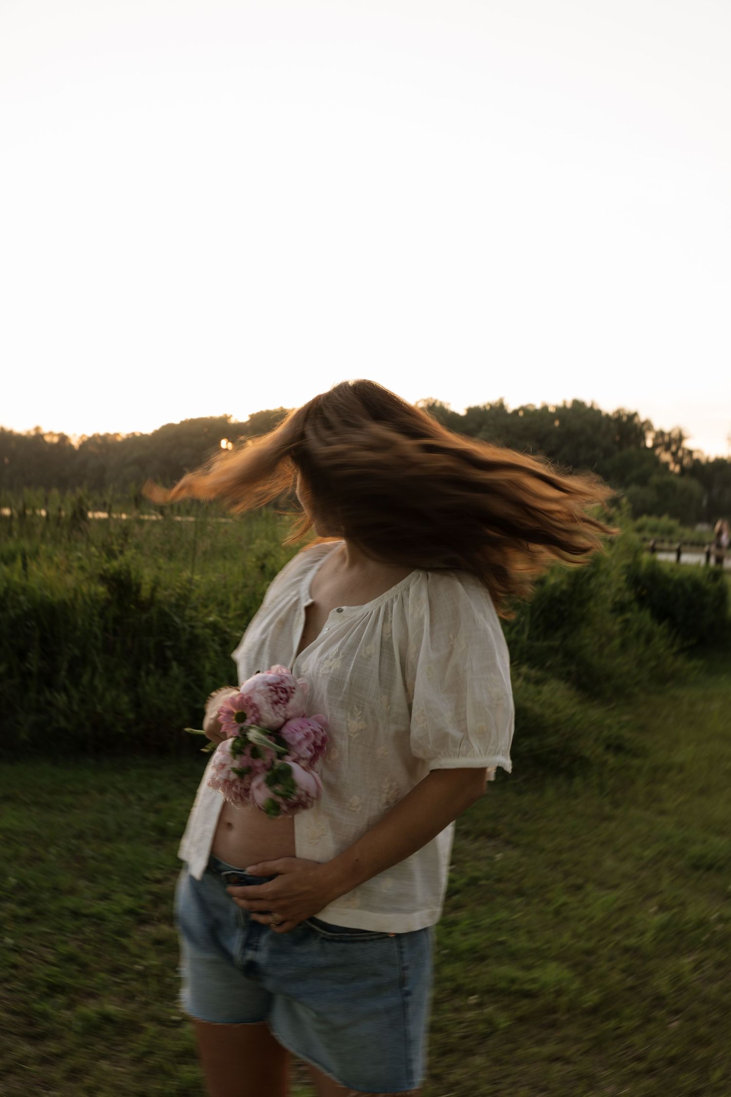 Pregnant woman in white top and denim shorts holding pink flowers, hair blowing in wind at sunset in a park.