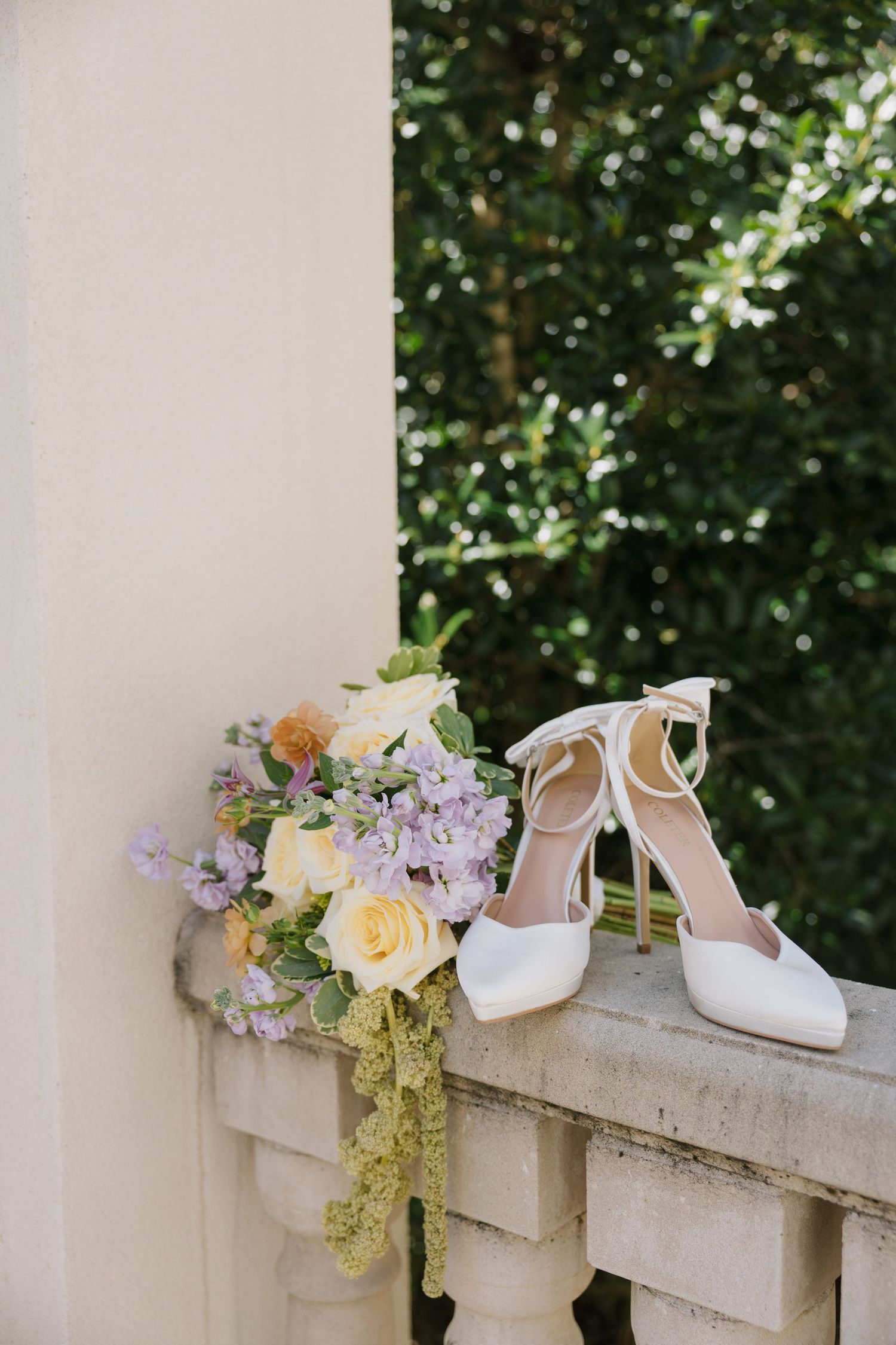 Bridal bouquet of cream roses and lavender flowers beside white ankle-strap heels on a stone balustrade.