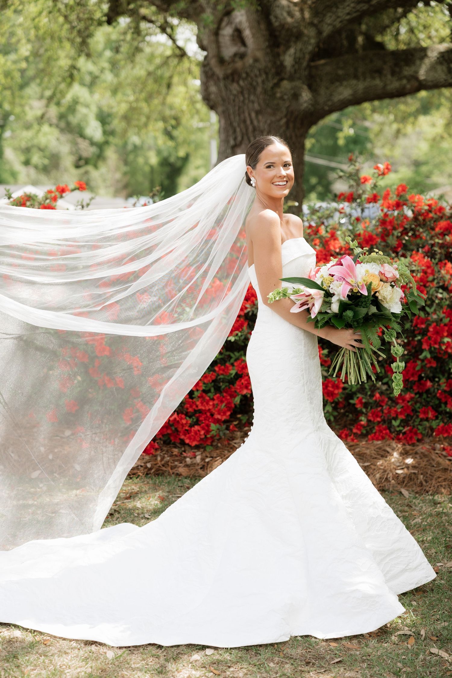 Bride in strapless lace mermaid gown with flowing veil holding bouquet, posing before red flowers.
