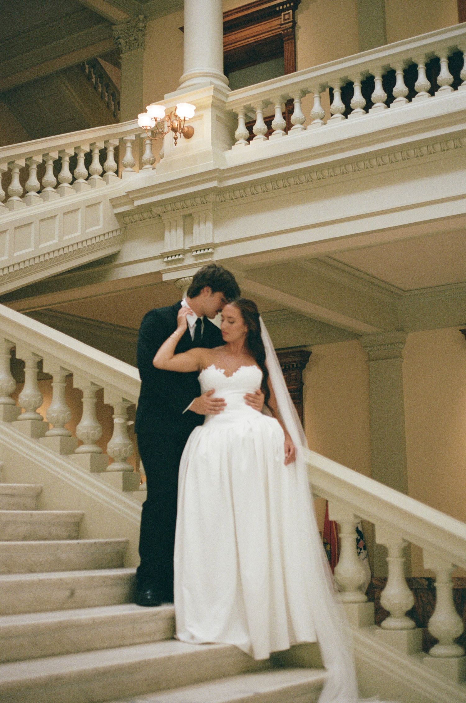 Bride in a strapless white gown and long veil embracing her groom in a black suit on an elegant marble staircase.