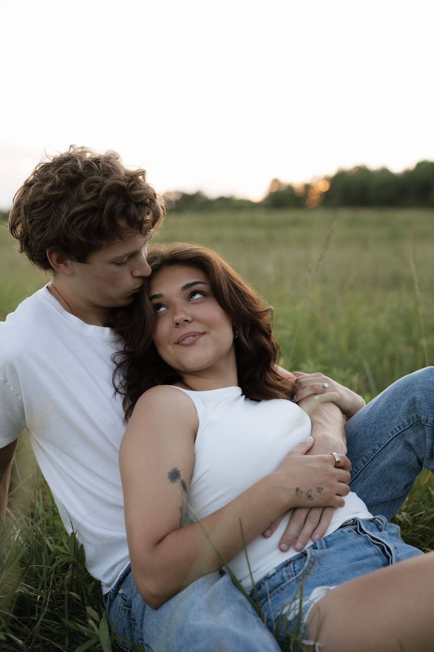 A couple sits in a green field at sunset, embracing tenderly in white tops and jeans.