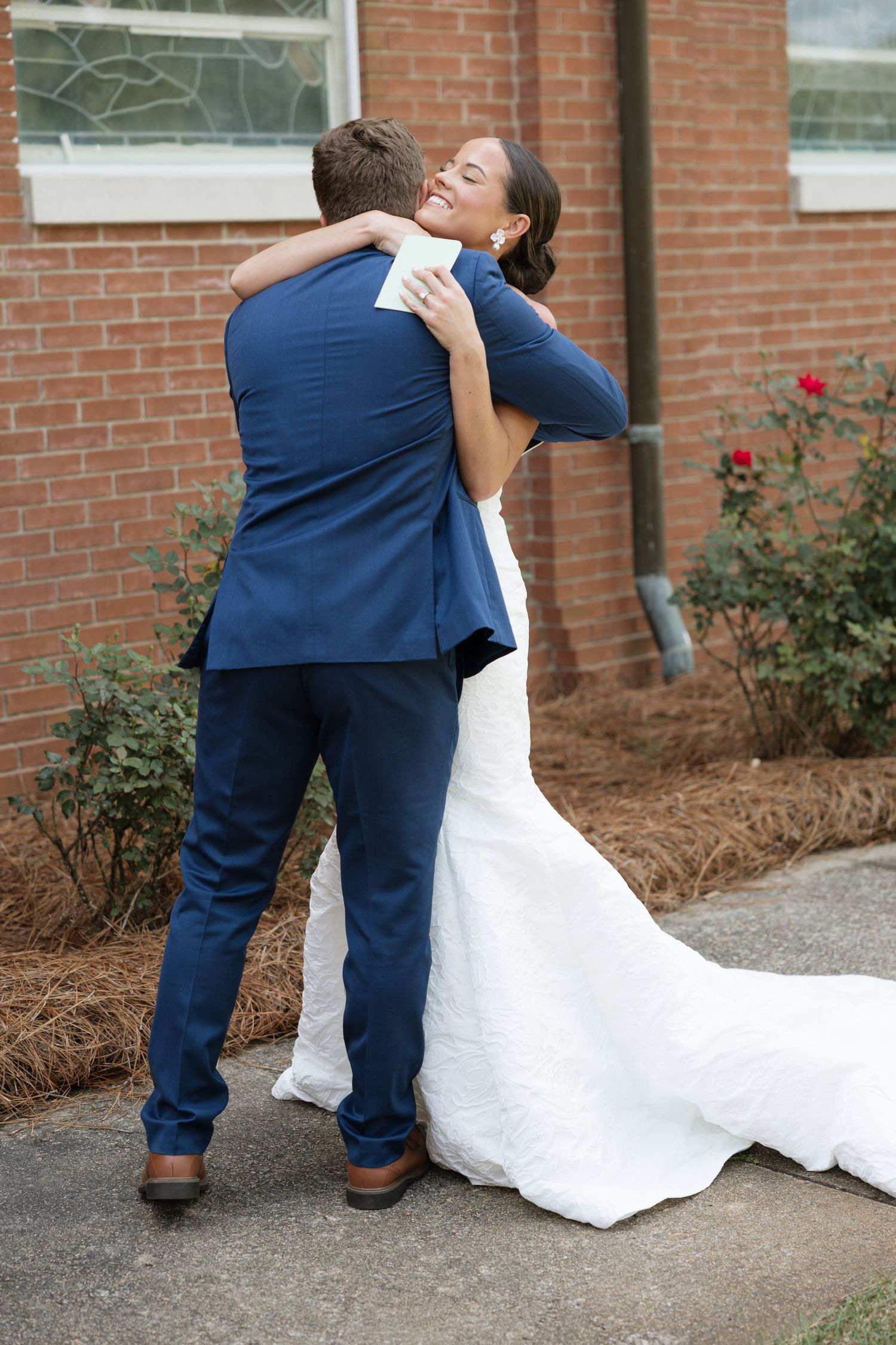 Bride in white gown hugging groom in navy suit outside brick church on wedding day.