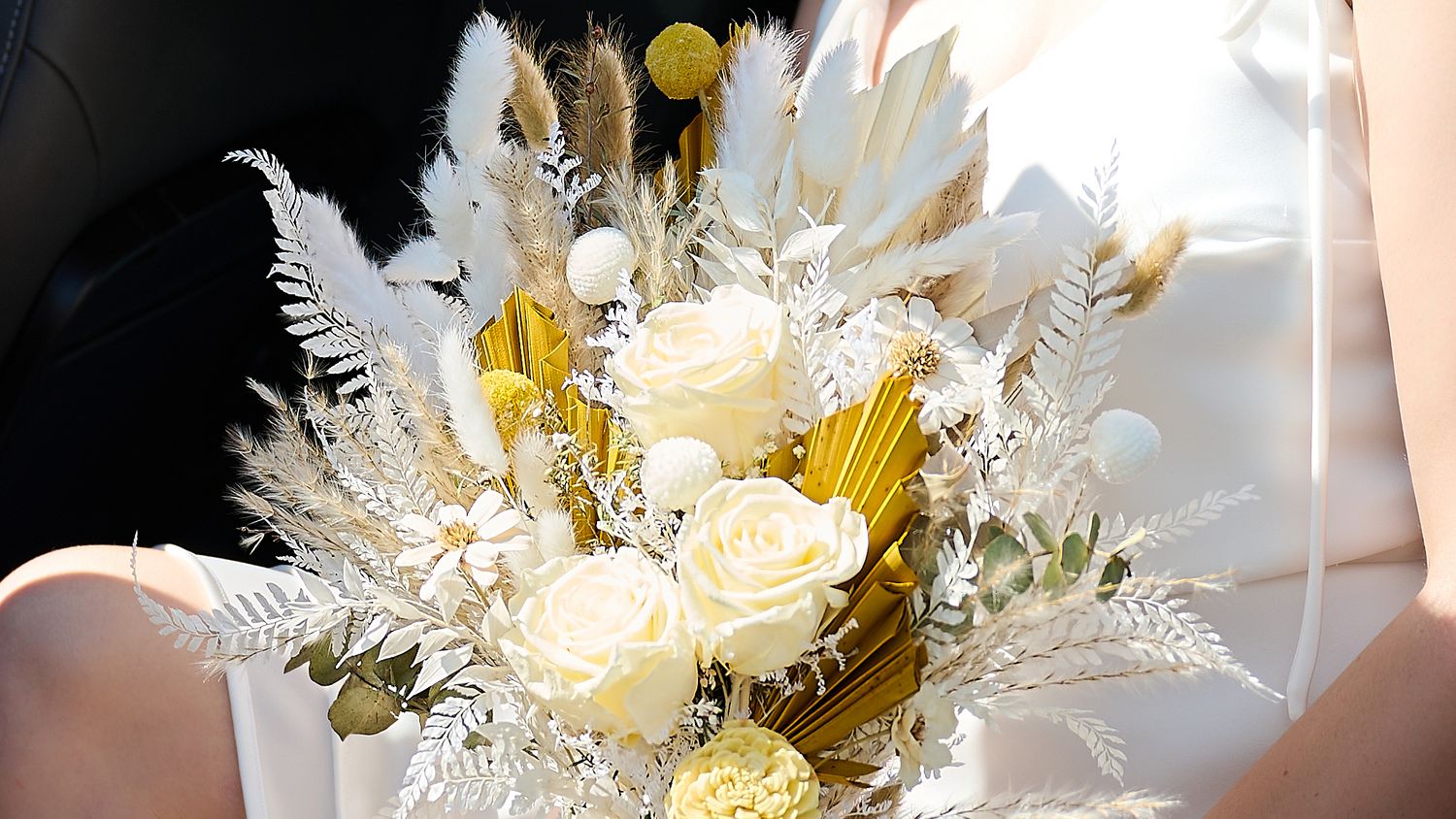 A bride holding a stunning white and gold bouquet with dried flowers and roses on her wedding day.