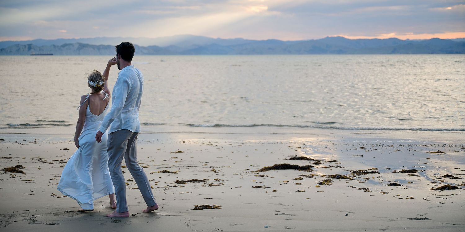 Couple walks barefoot along a peaceful beach at sunset with mountains in the background.