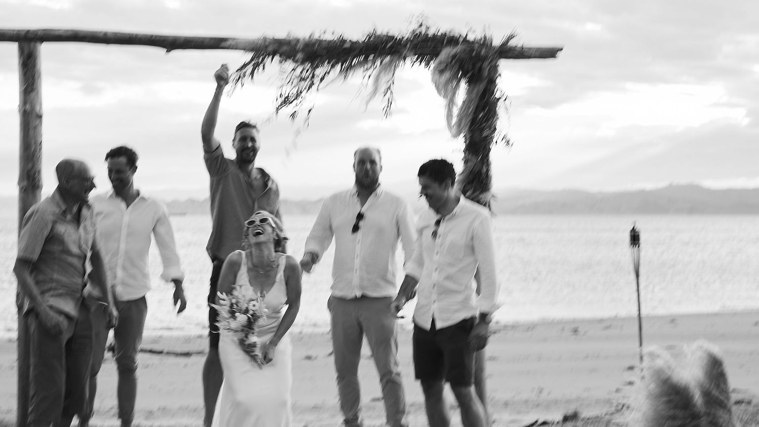Black and white photo of wedding party celebrating under rustic beach arch.