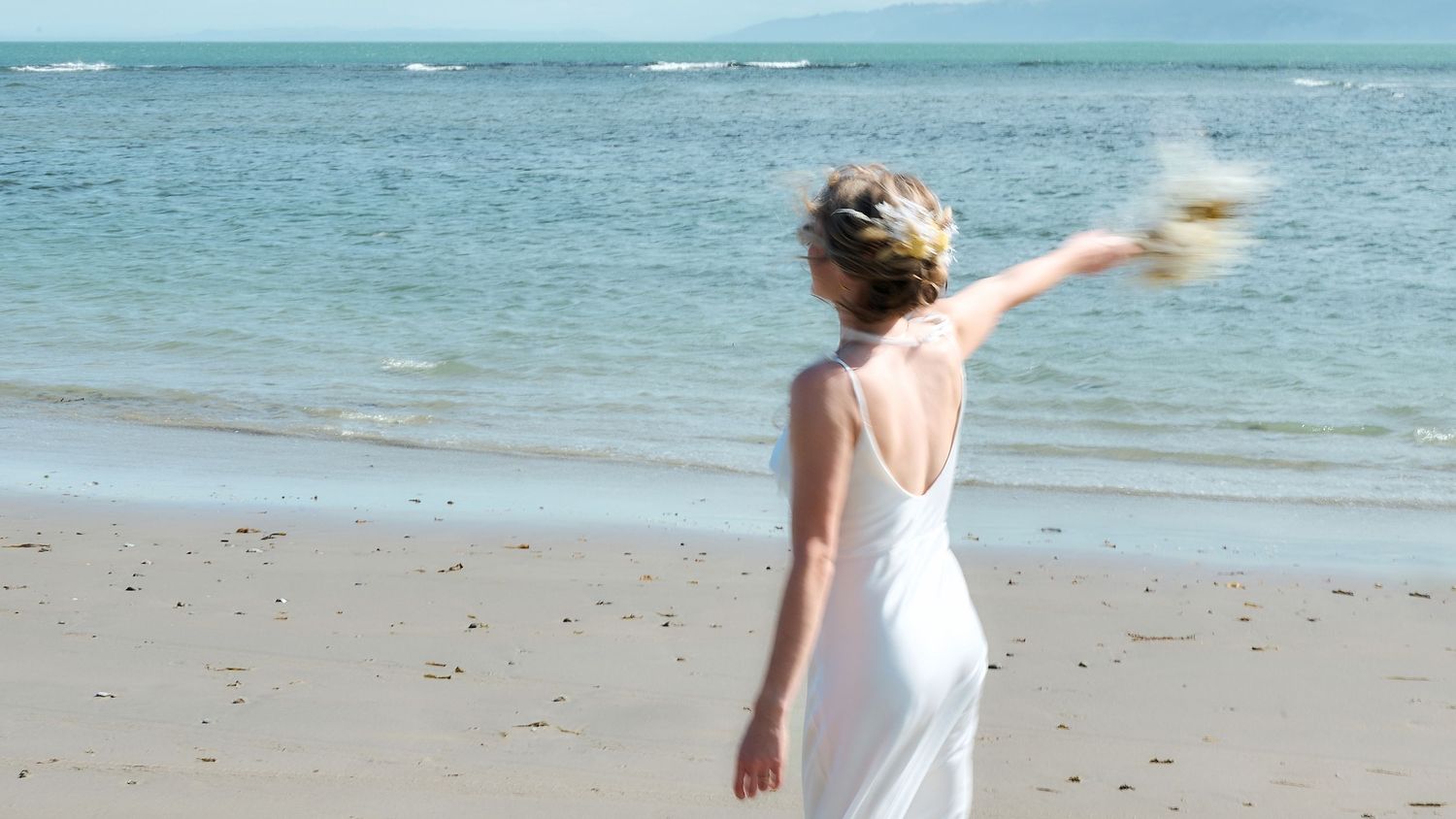 A bride in a white backless gown tossing her bouquet toward the ocean on a sunny beach.