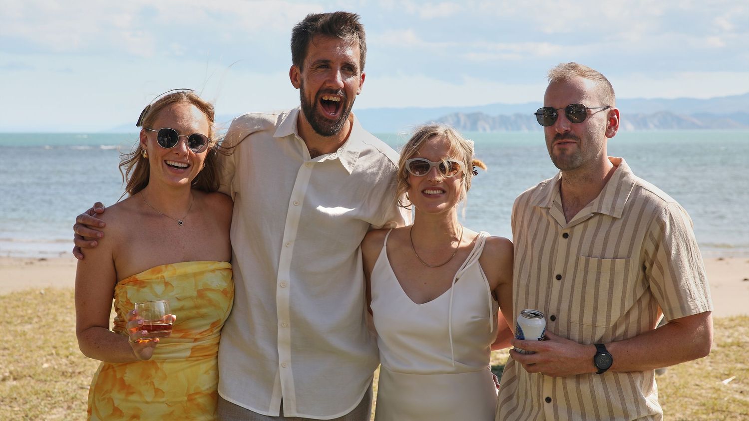 Four smiling friends pose together at outdoor beach wedding reception on a sunny day.