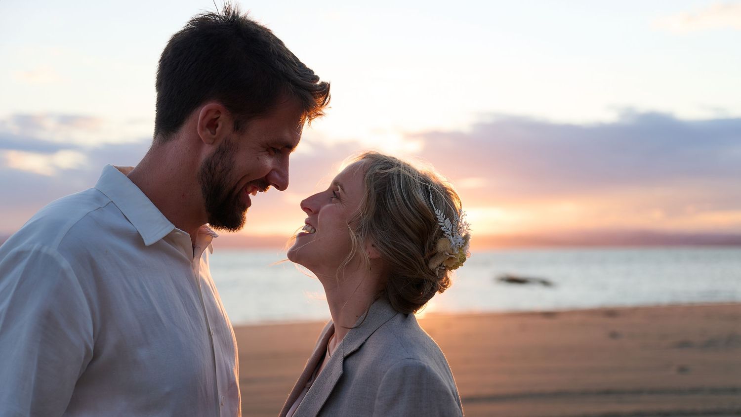 Close-up of a smiling couple facing each other on a beach during a pastel sunset.