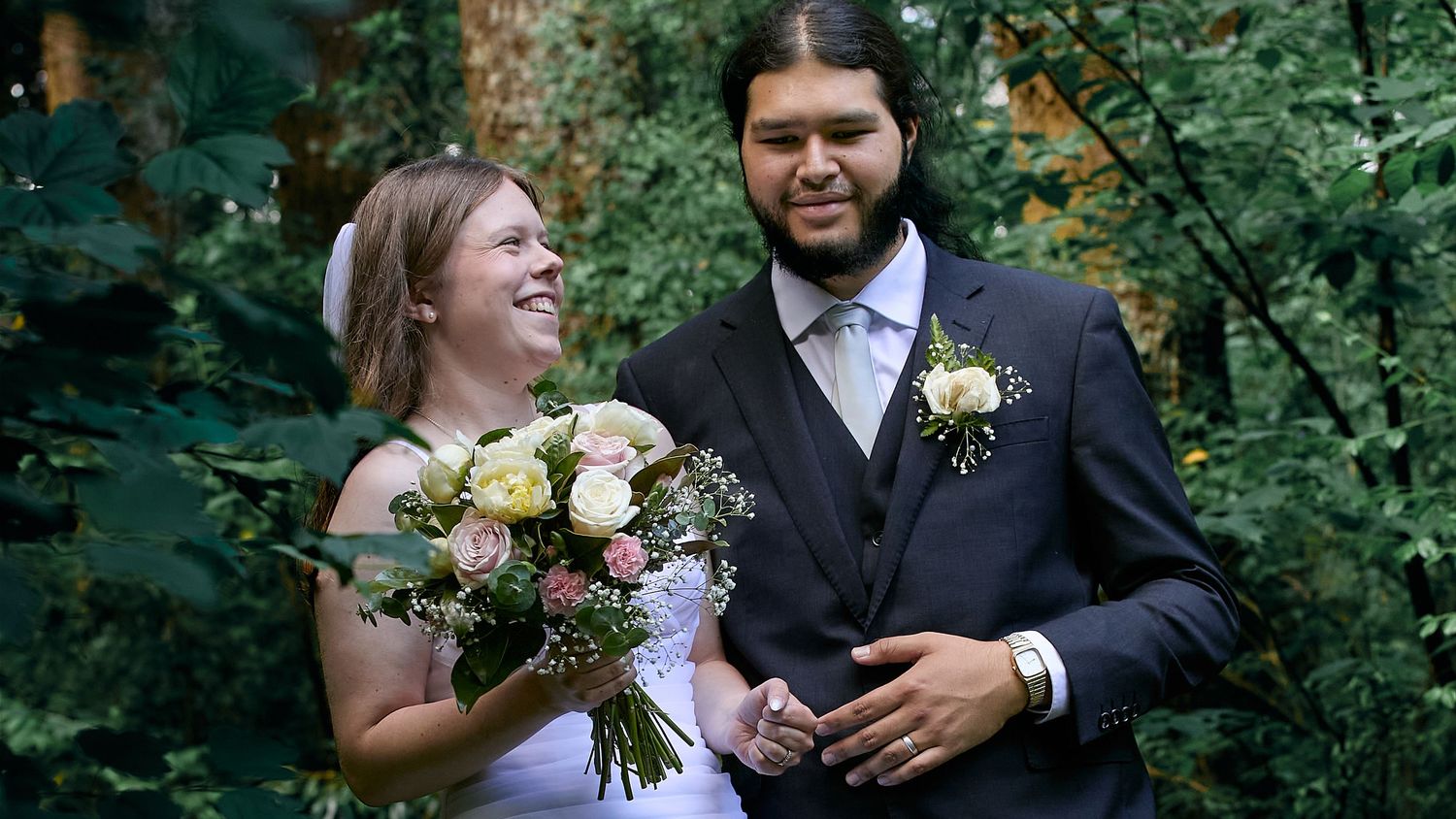 Smiling bride holding bouquet of pink and white roses standing with groom in suit in garden.