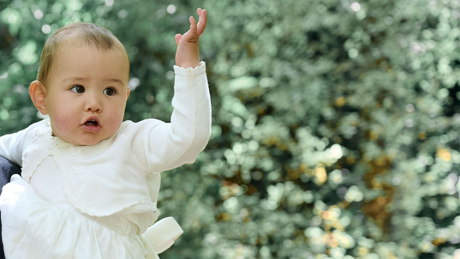 Adorable baby in a white dress raising one hand while being held outdoors near greenery.