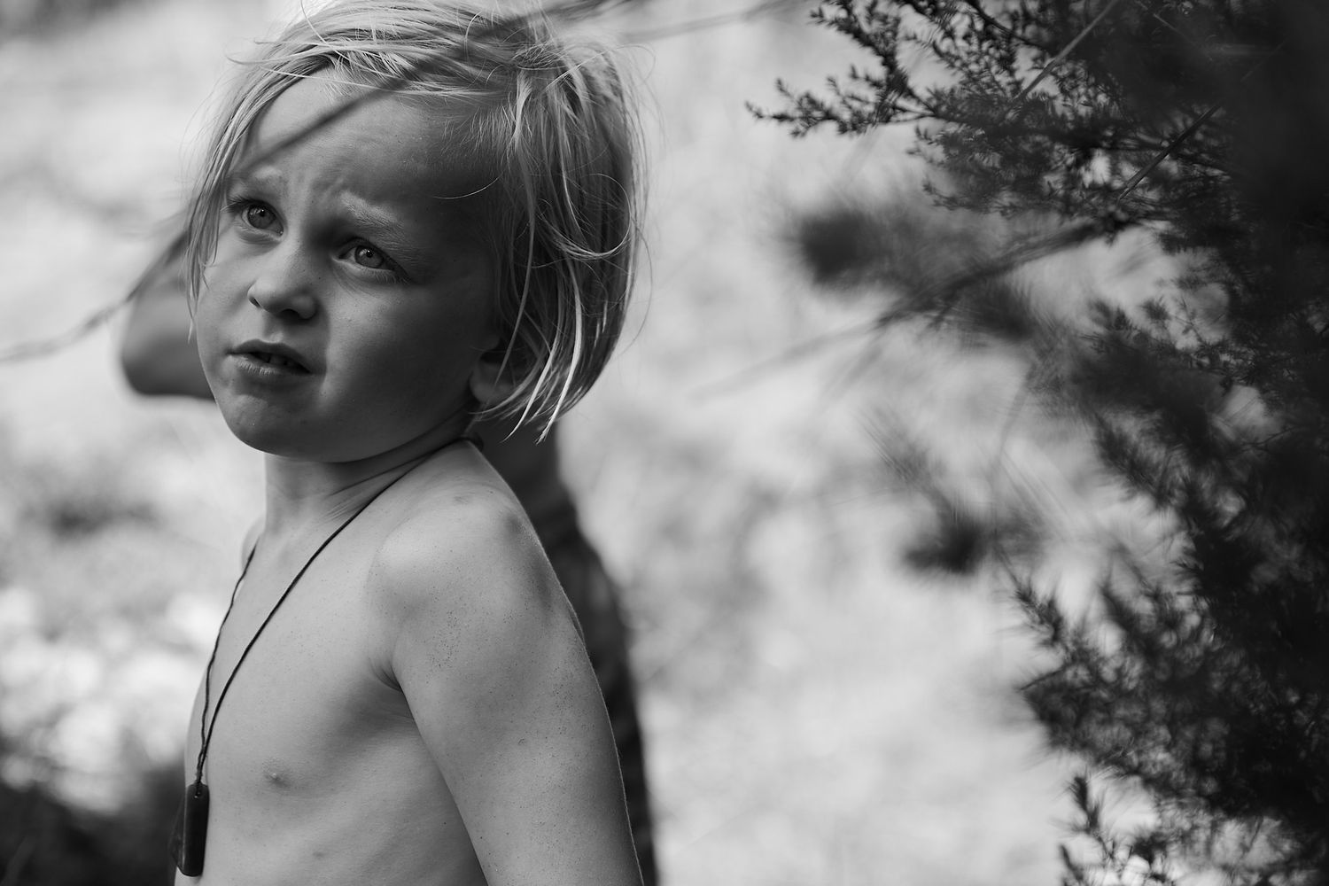 Black and white portrait of a young blonde child standing near pine trees outdoors.