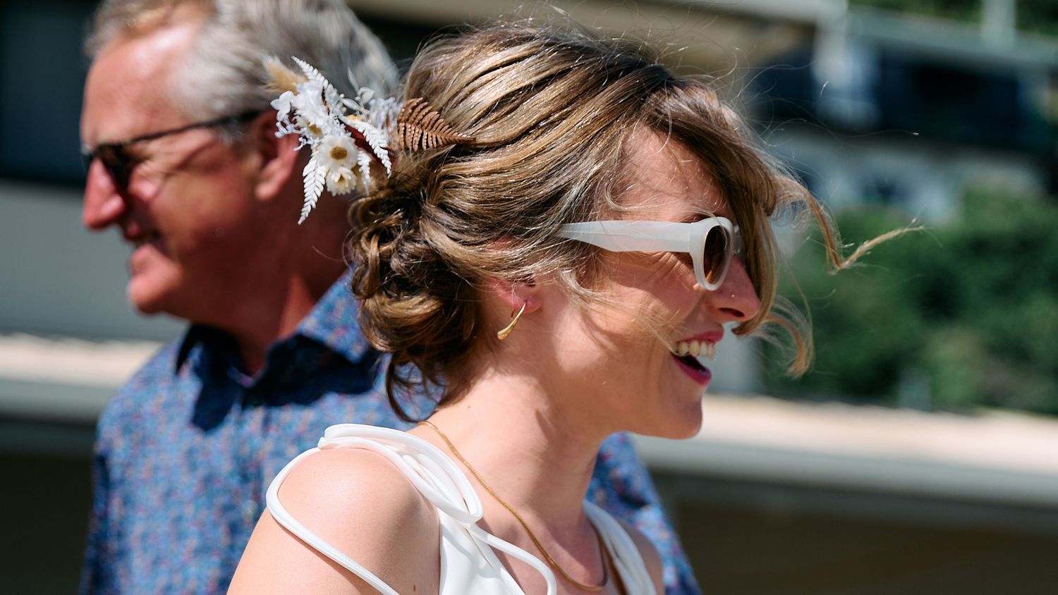 A smiling bride with a floral hair accessory and white sunglasses walks outdoors on her wedding day.