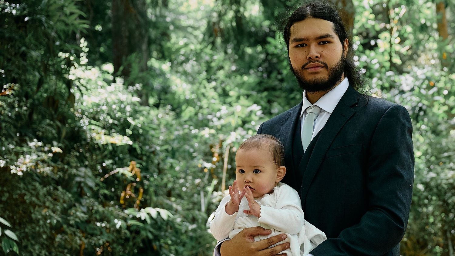 Young bearded man in a navy suit holding a baby in white clothing in a green garden.