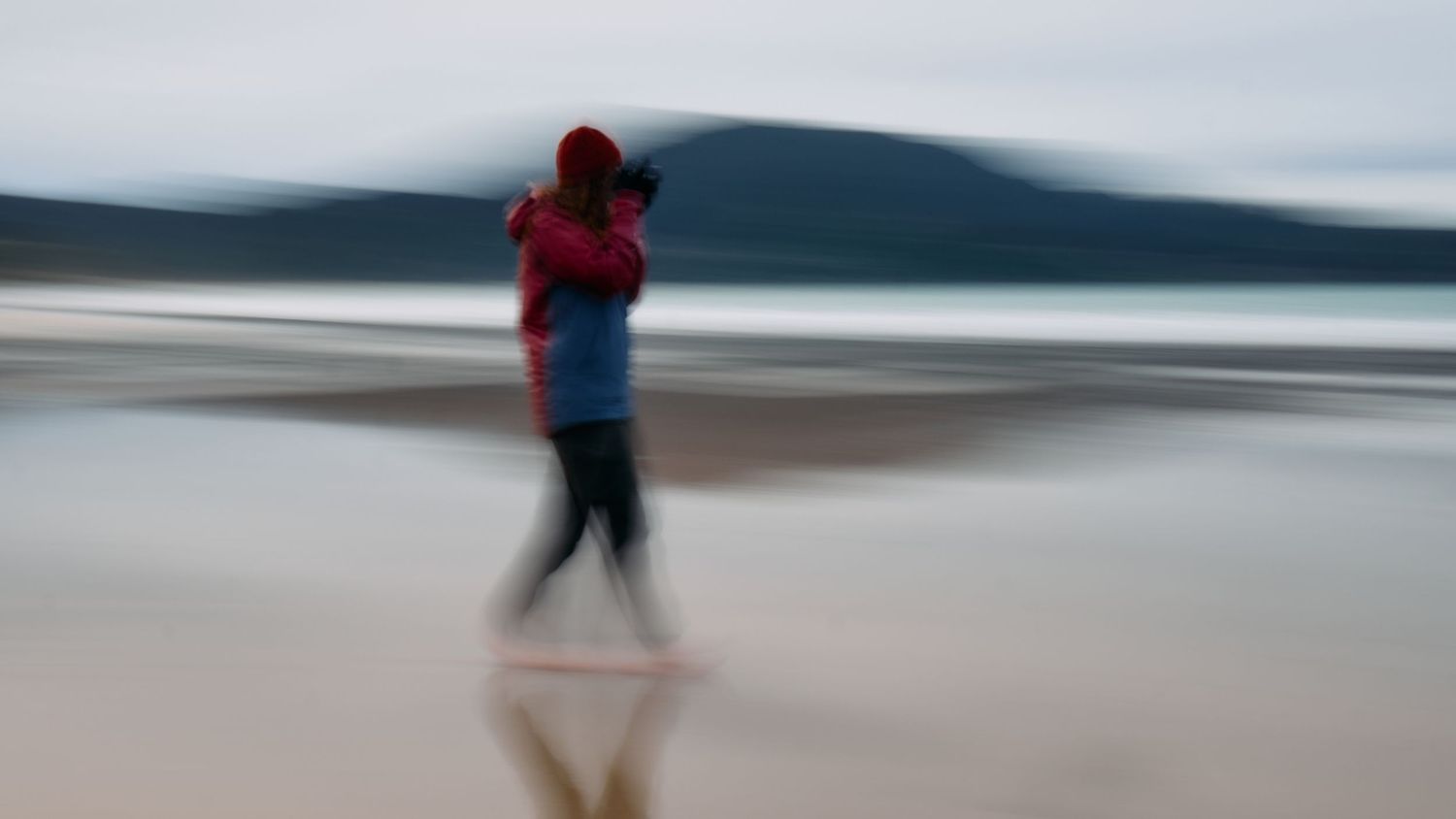 Motion blur photo of person carrying child on reflective beach with misty mountain background.