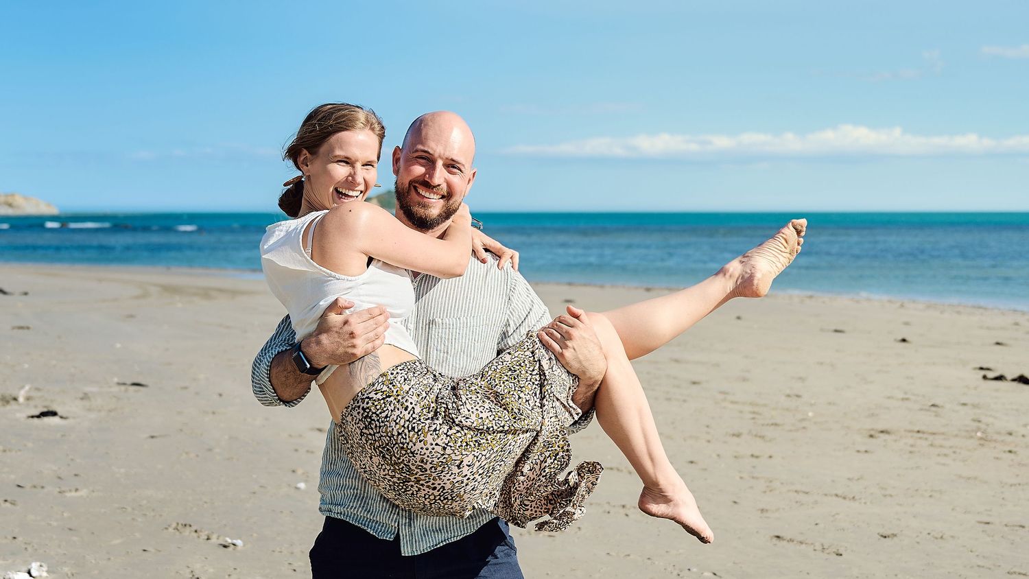 Groom carries bride on beach with blue ocean and sky in background.