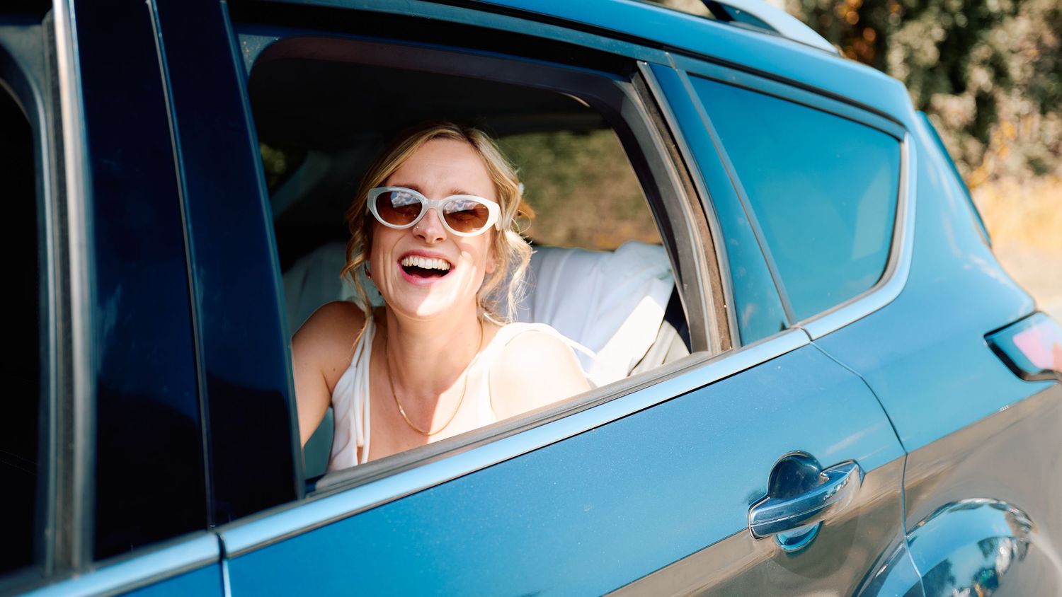 A happy bride in sunglasses laughing while leaning out of a blue car window on her wedding day.