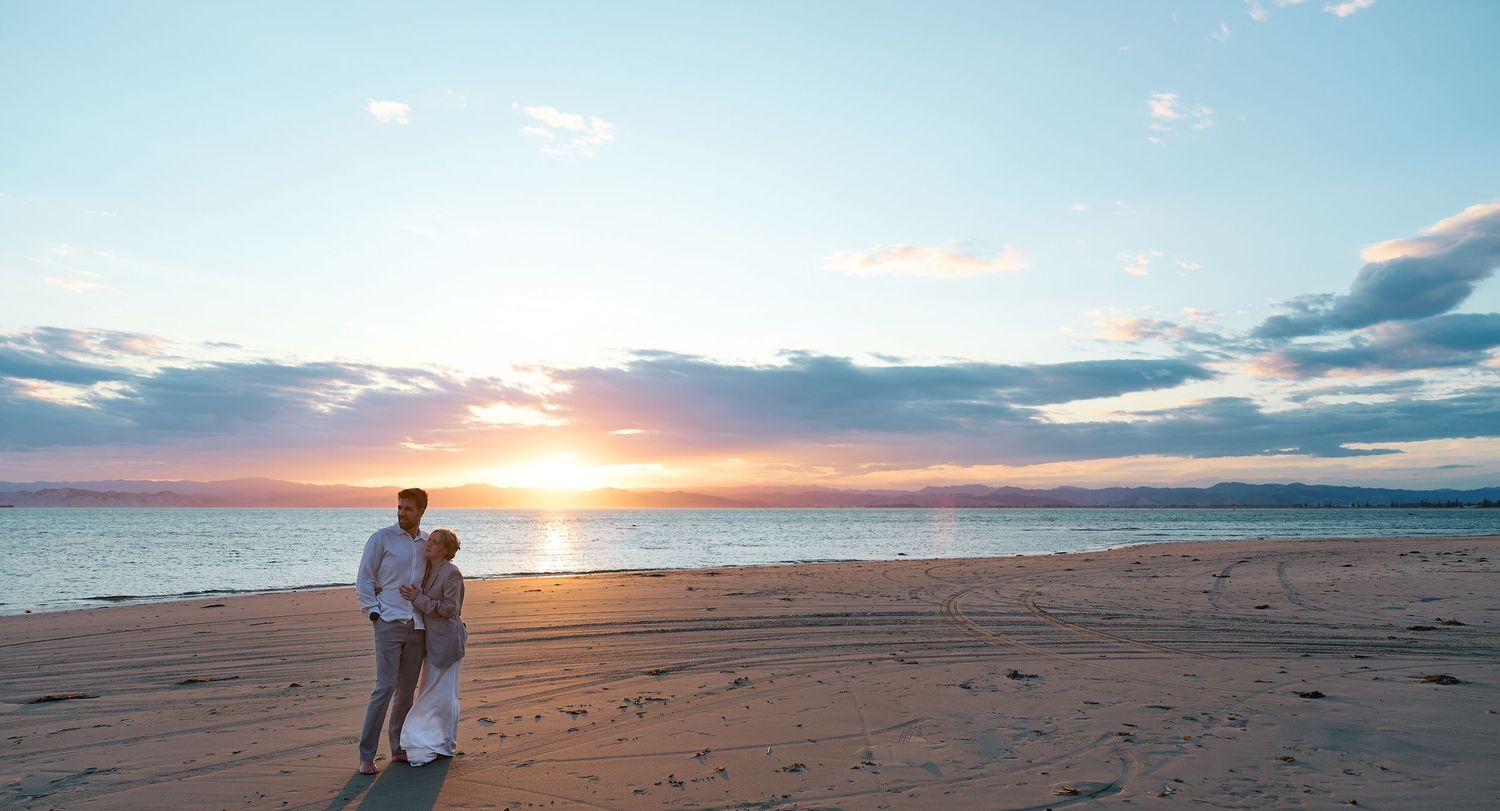 Couple stands together on a sandy beach watching a vibrant golden sunset over the ocean.