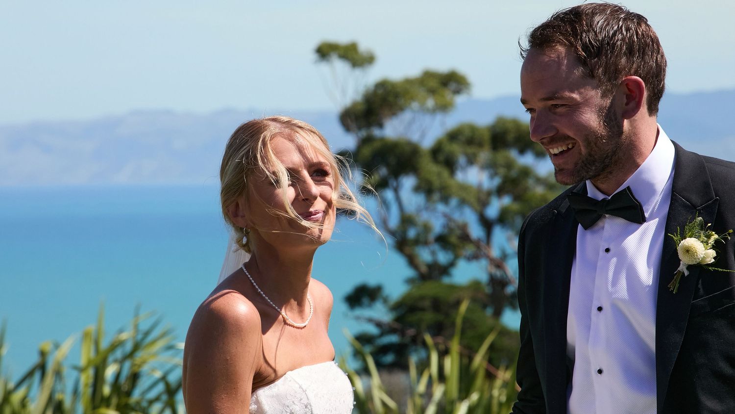 A smiling bride and groom share a joyful moment outdoors with a stunning blue ocean backdrop.