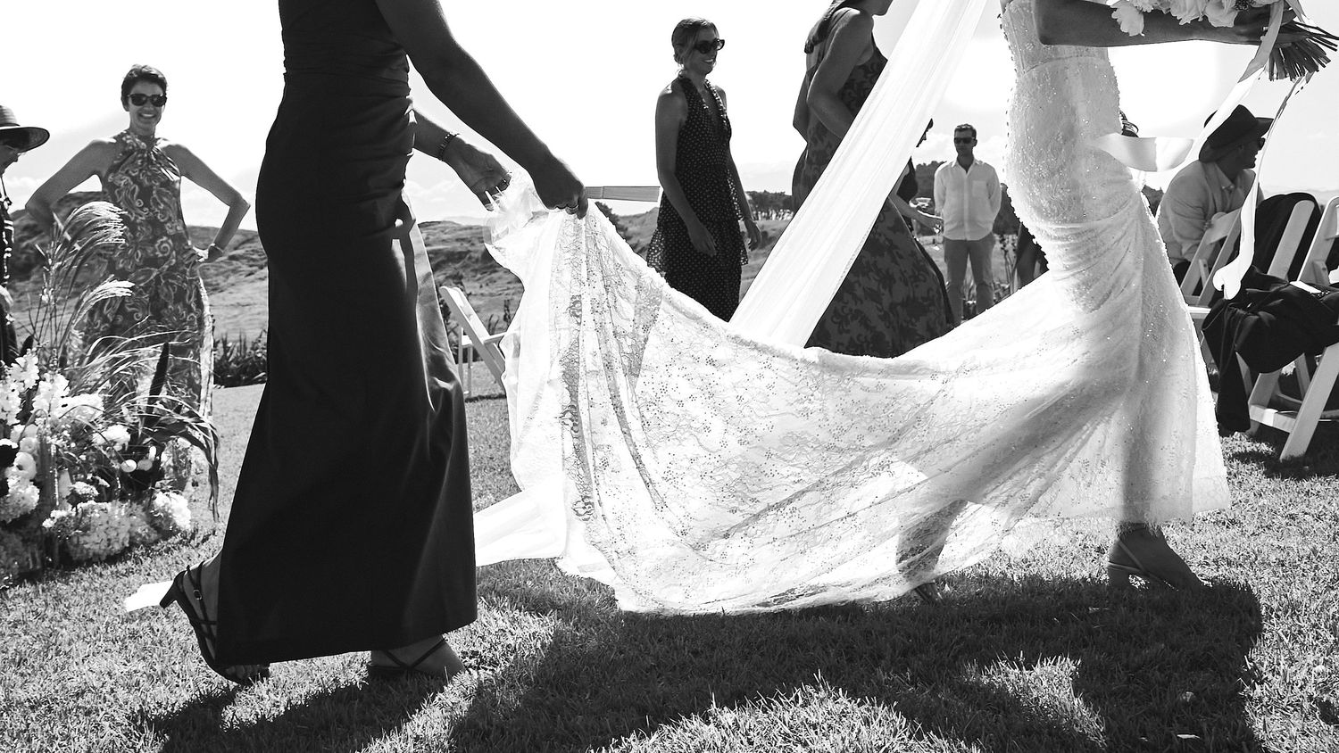 Black and white photo of bridesmaids holding bride's lace wedding dress train during outdoor ceremony.