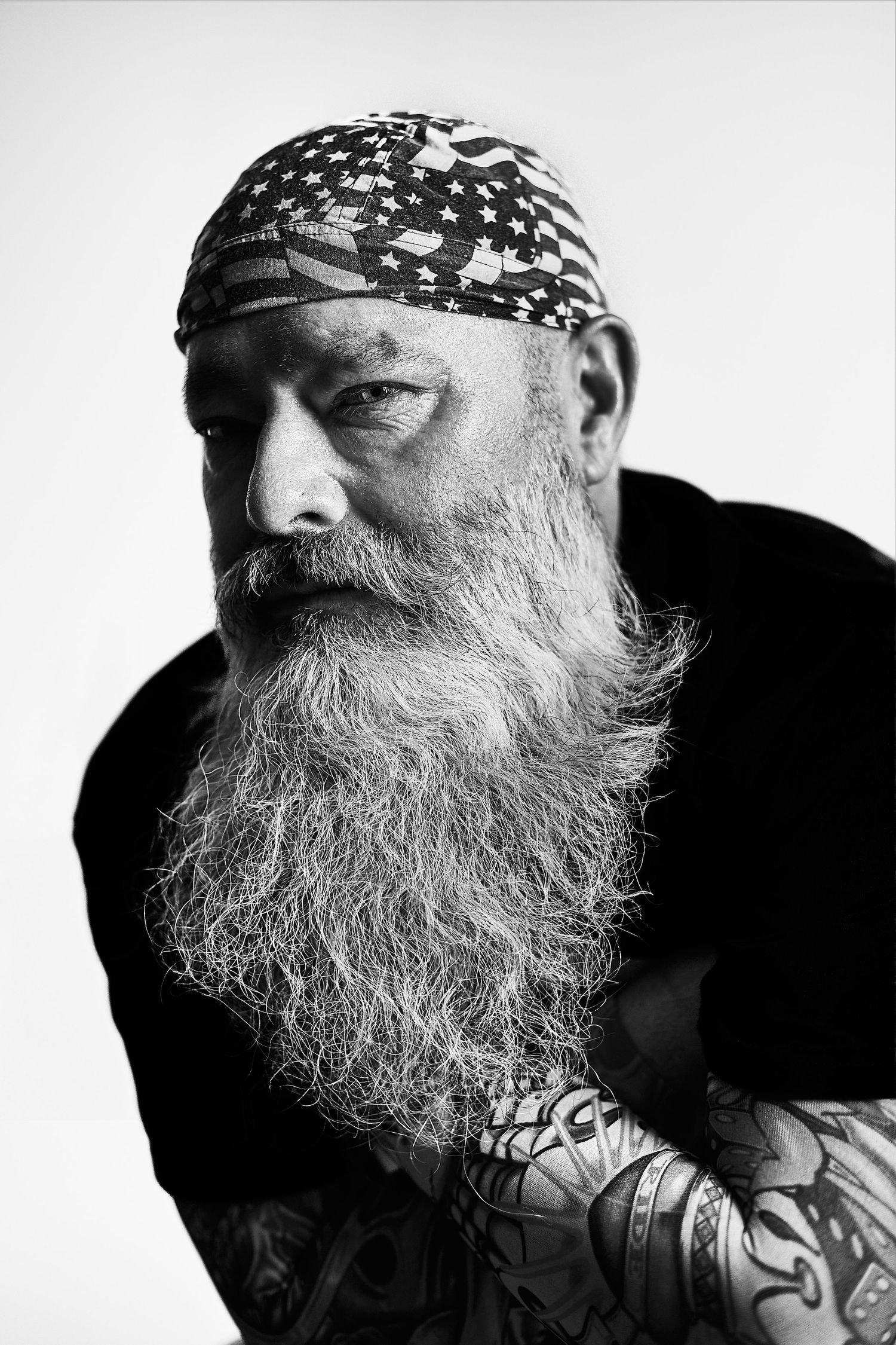Black and white portrait of a tattooed older man with a long gray beard wearing a star-patterned bandana.