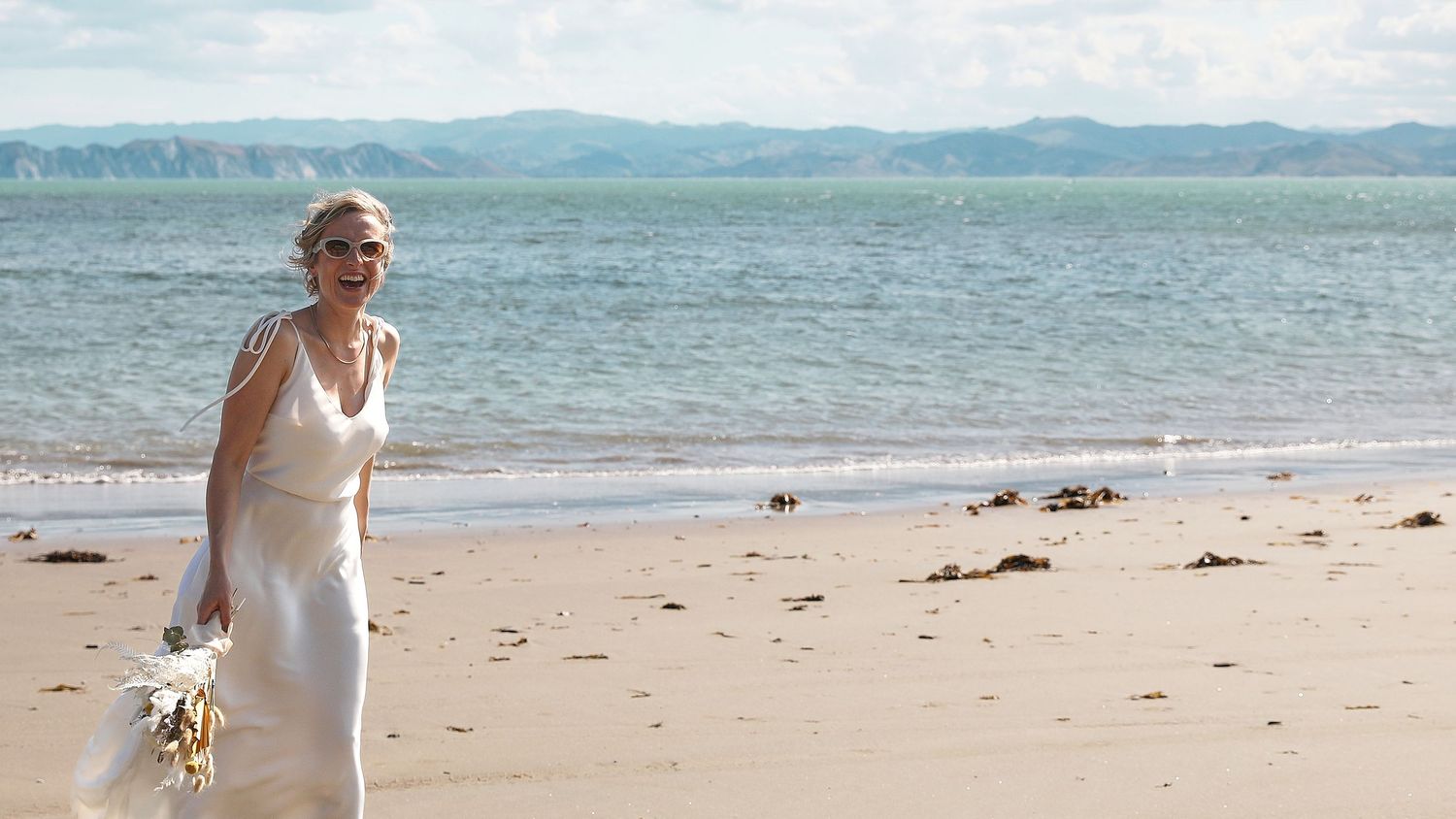 Elegant bride in white gown walks along scenic beach with bouquet.