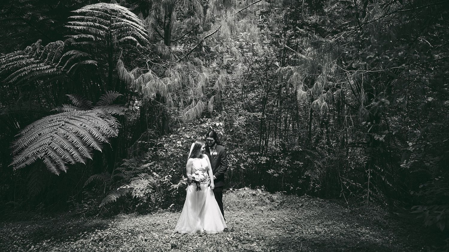 Black and white photo of a bride and groom embracing in a lush forest setting with large ferns.