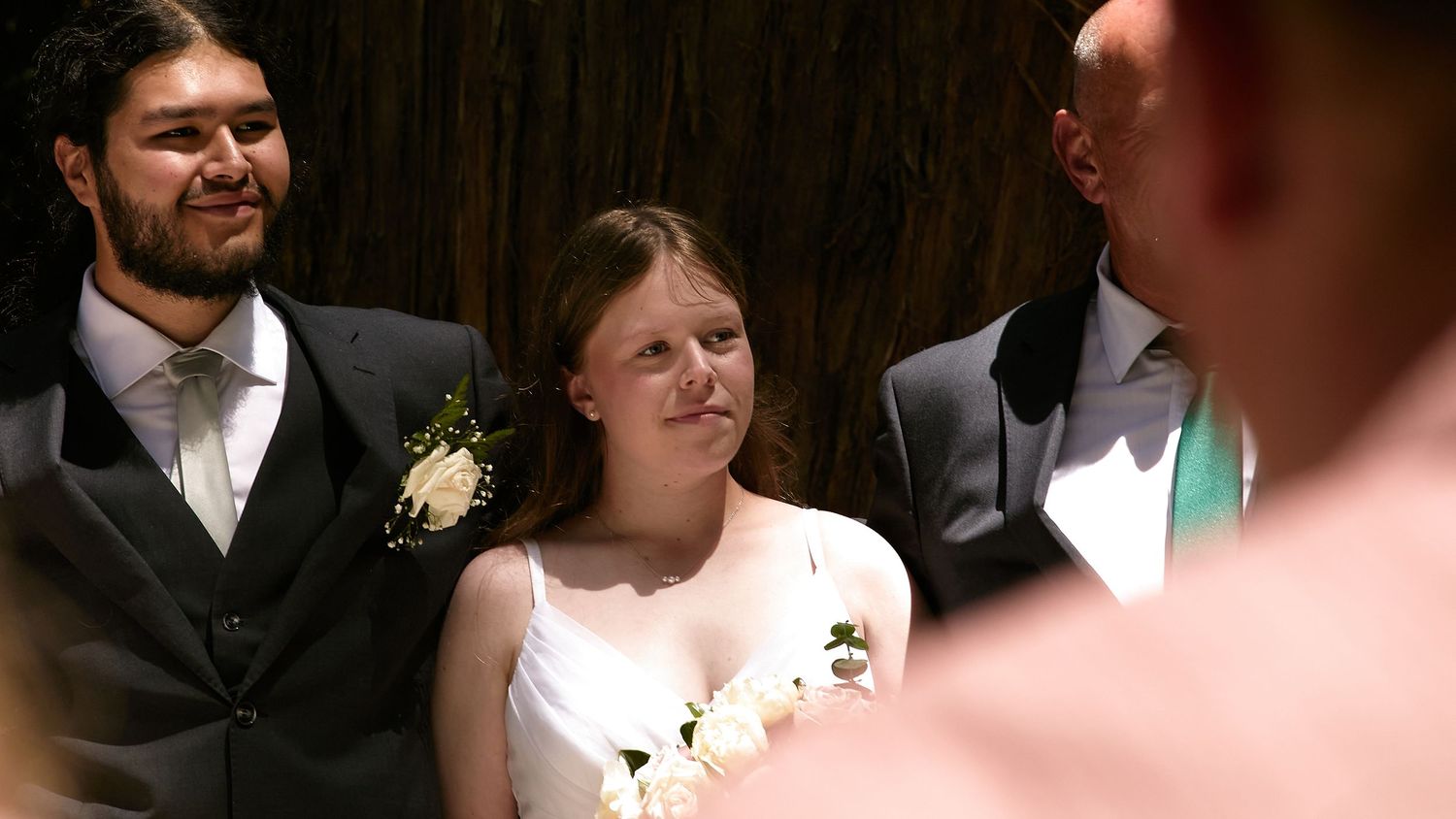 Bride in white dress smiling during outdoor forest wedding ceremony with groom in dark suit beside her.