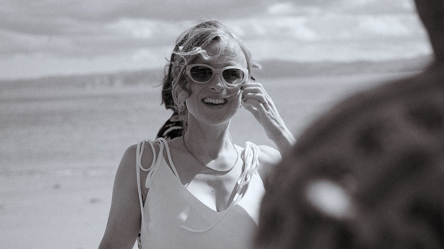 A joyful bride in white sunglasses smiles on a beach in a candid black and white wedding photo.