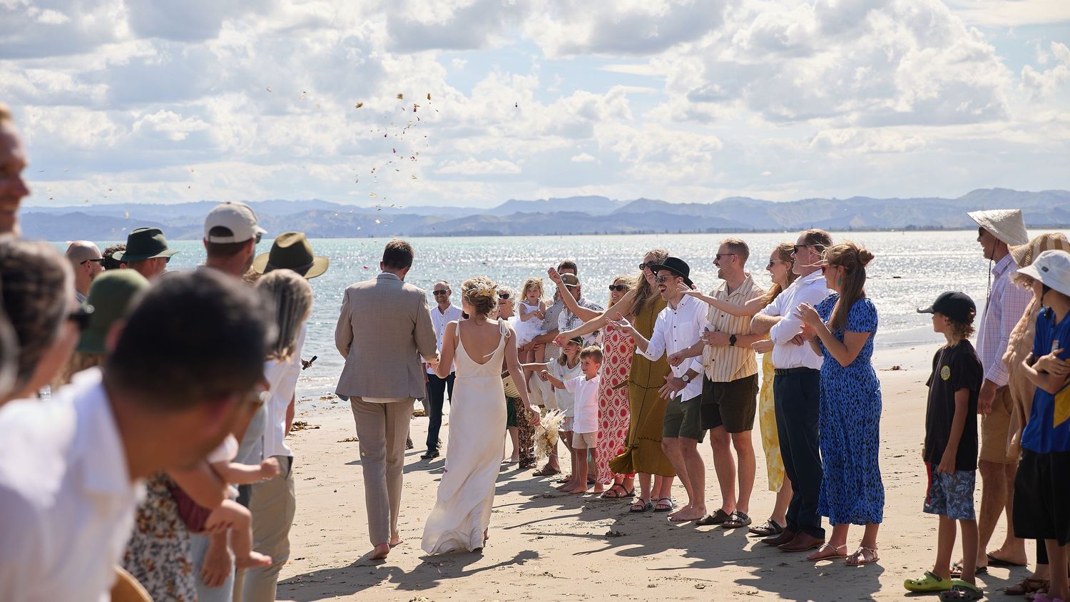 Bride and groom walk through confetti tunnel of cheering wedding guests on sunny beach ceremony.