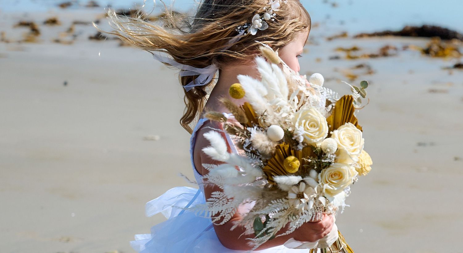 Young girl in a white dress holding a bouquet of dried flowers on a windy beach with hair blowing.