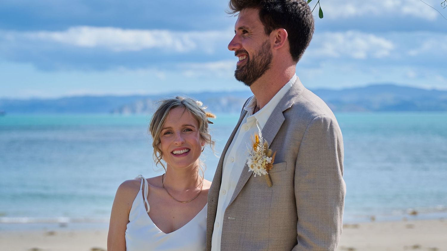 Bride and groom smiling together on a sunny beach with blue ocean and sky in the background.