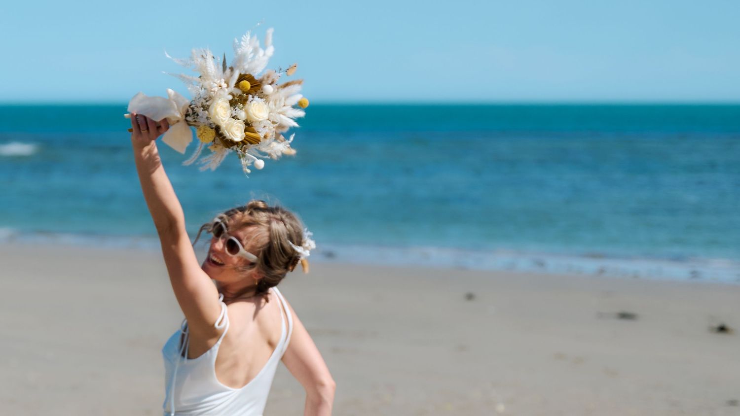 Joyful bride in sunglasses raises bouquet high on sunny beach wedding day.