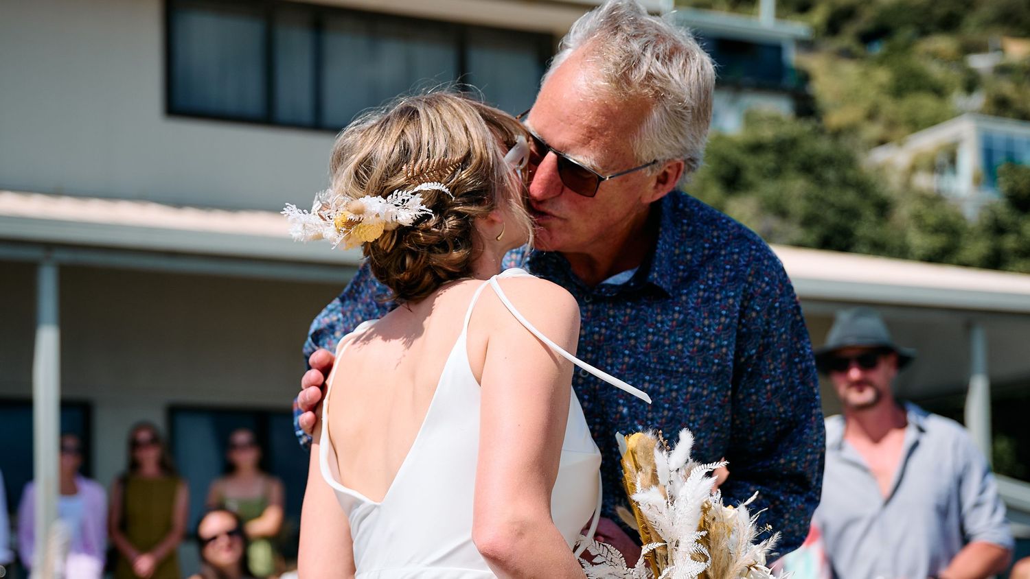 An older gentleman shares a warm embrace with a bride holding a dried flower bouquet at a wedding.
