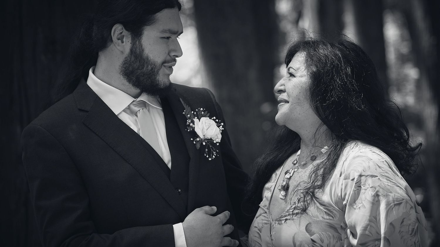 Black and white photo of groom in suit with boutonniere smiling at woman in floral dress at wedding.